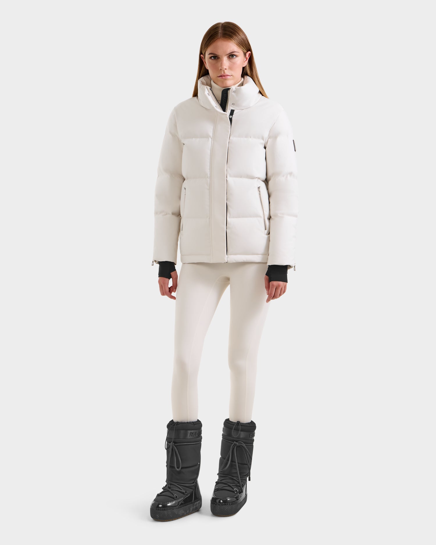 A woman faces forward wearing the Rudsak JOON H - 8125586 Vanilla women’s down jacket with cream leggings and black winter boots against a plain light background—a true cold-weather essential.
