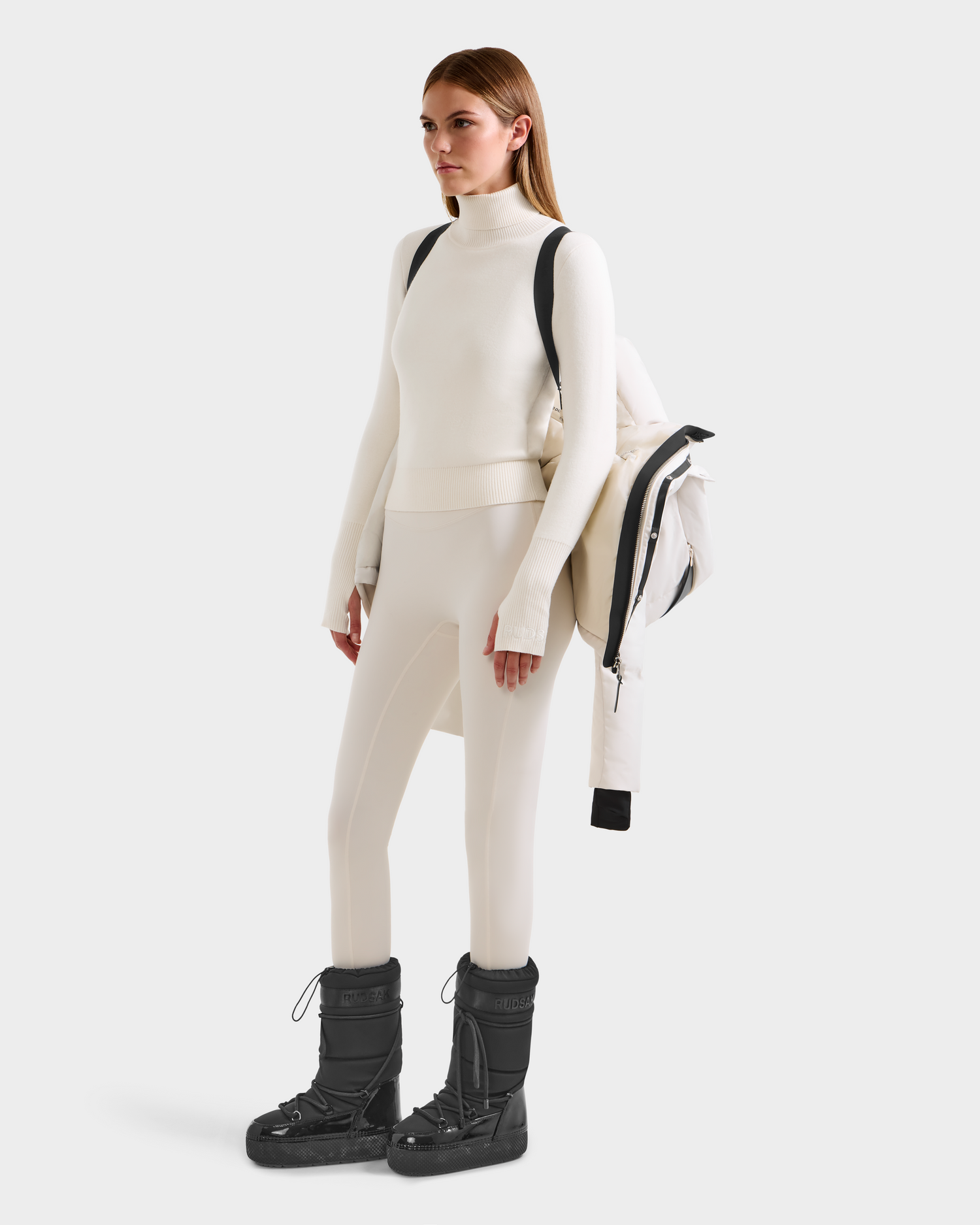 A woman in a fitted white turtleneck, matching leggings, and black snow boots stands holding the Rudsak JOON H - 8125586 Vanilla women’s down jacket over her shoulder with a backpack strap, against a light background.