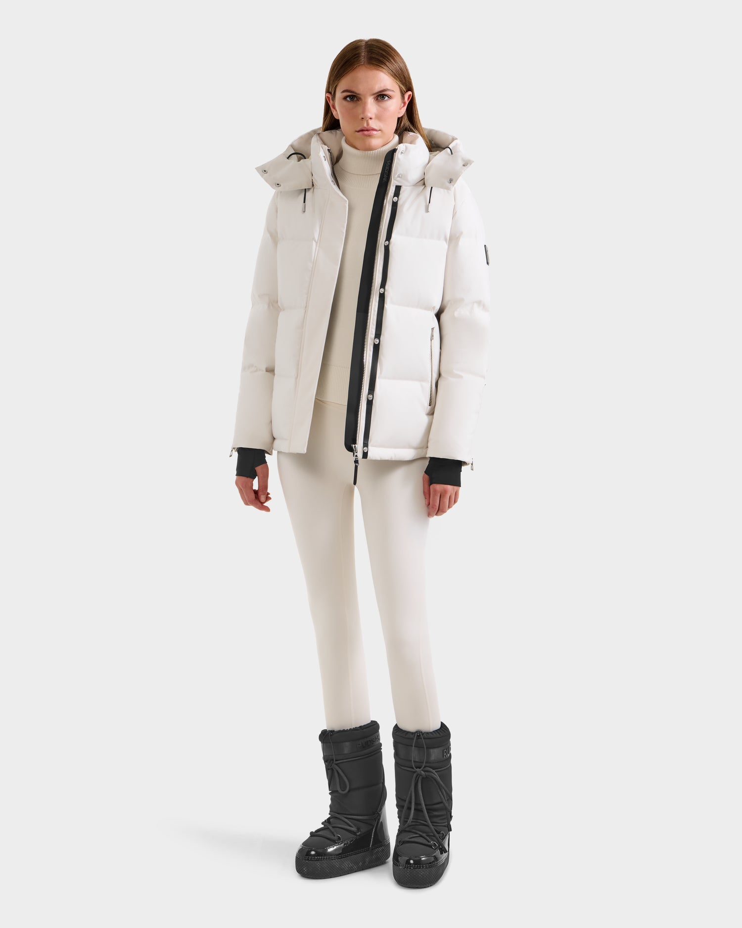 A person stands facing forward in the Rudsak JOON H - 8125586 Vanilla women’s down jacket, styled with a white turtleneck, white leggings, and black snow boots for an effortlessly stylish winter look against a plain light background.