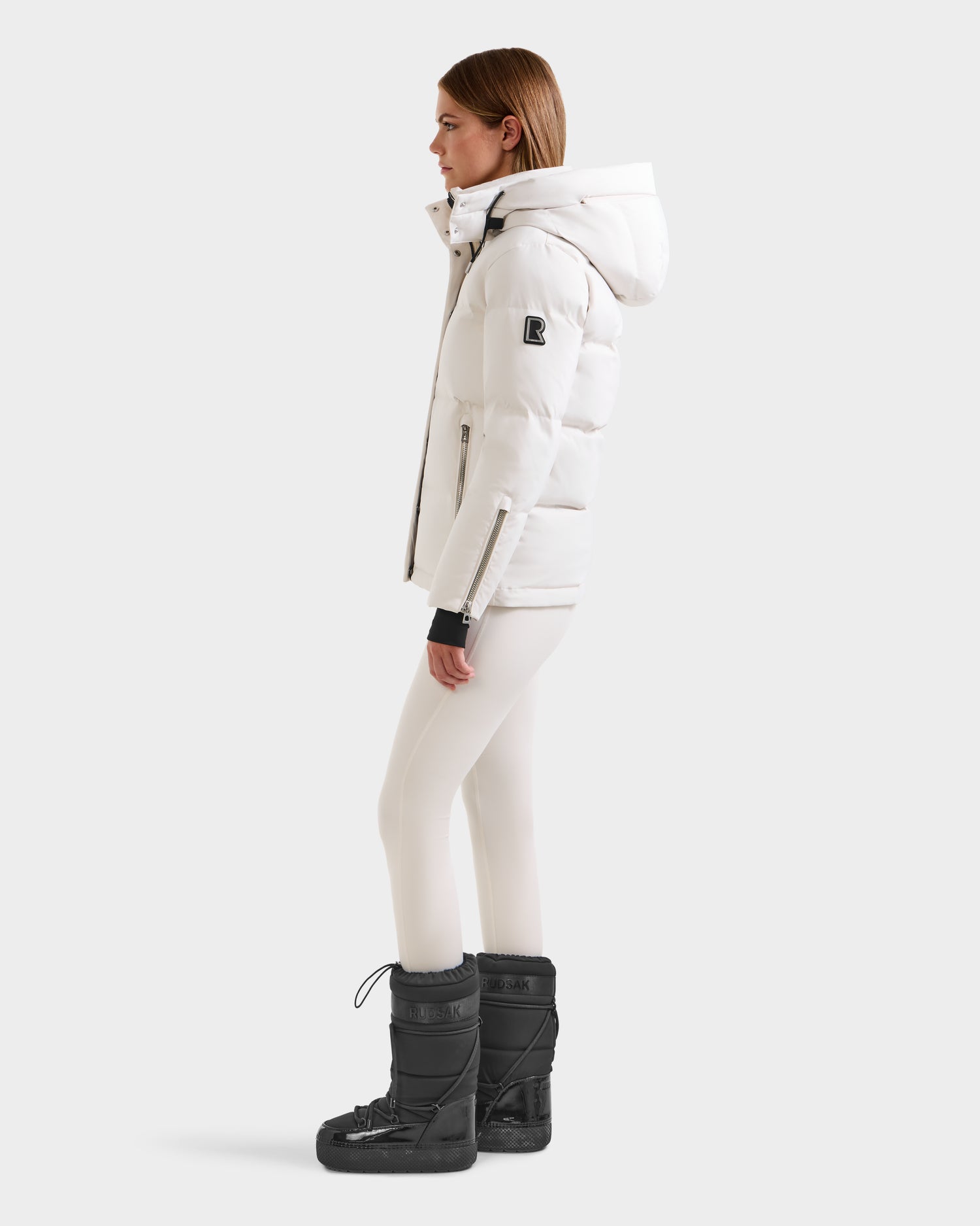 A woman in profile wears the Rudsak JOON H - 8125586 Vanilla women’s down jacket with a hood, paired with white leggings and black boots against a plain light gray background—perfect for cold weather.