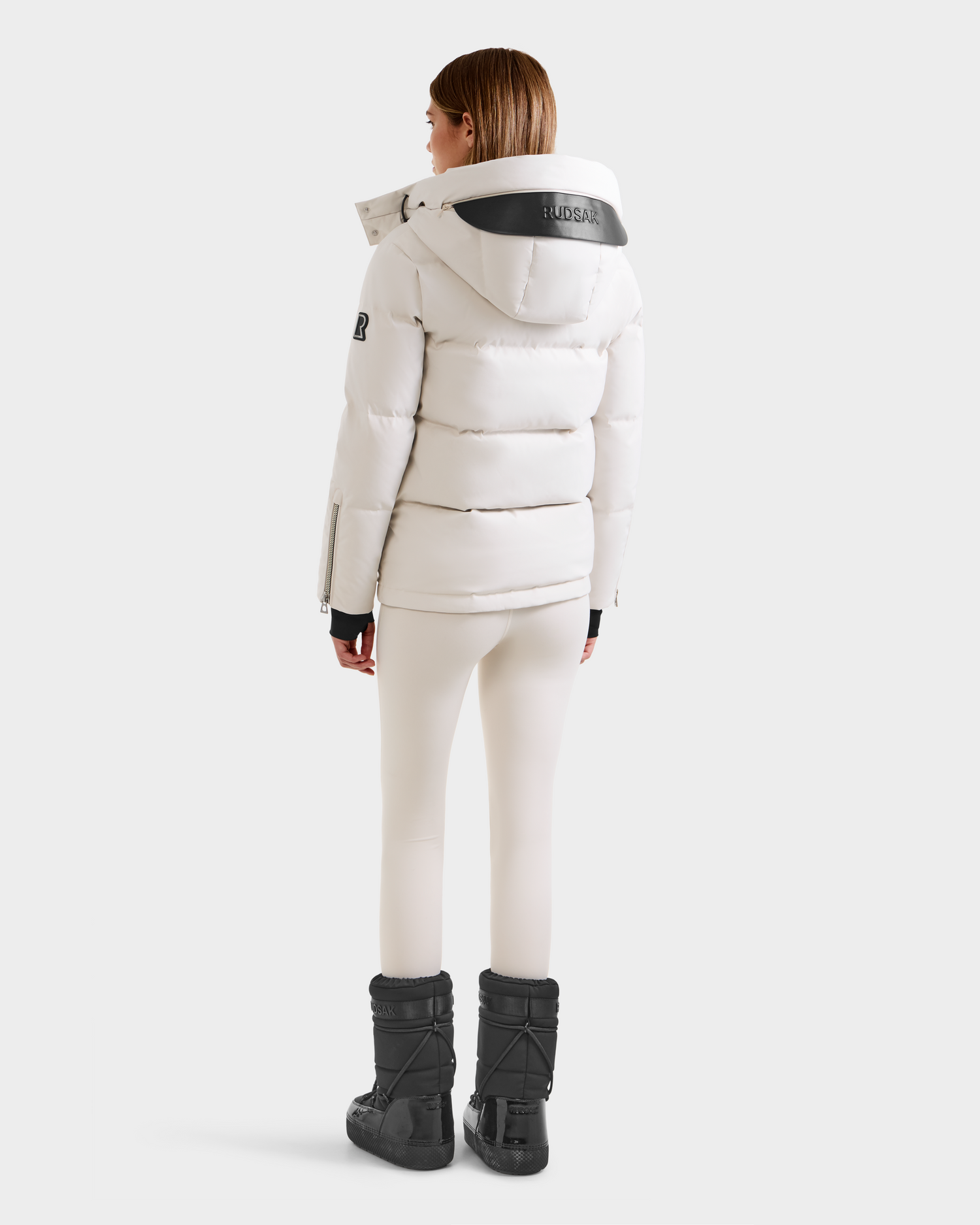 A woman faces away wearing the Rudsak JOON H - 8125586 Vanilla women’s down jacket with a hood, white leggings, and black boots against a plain light background—an effortless cold-weather essential.