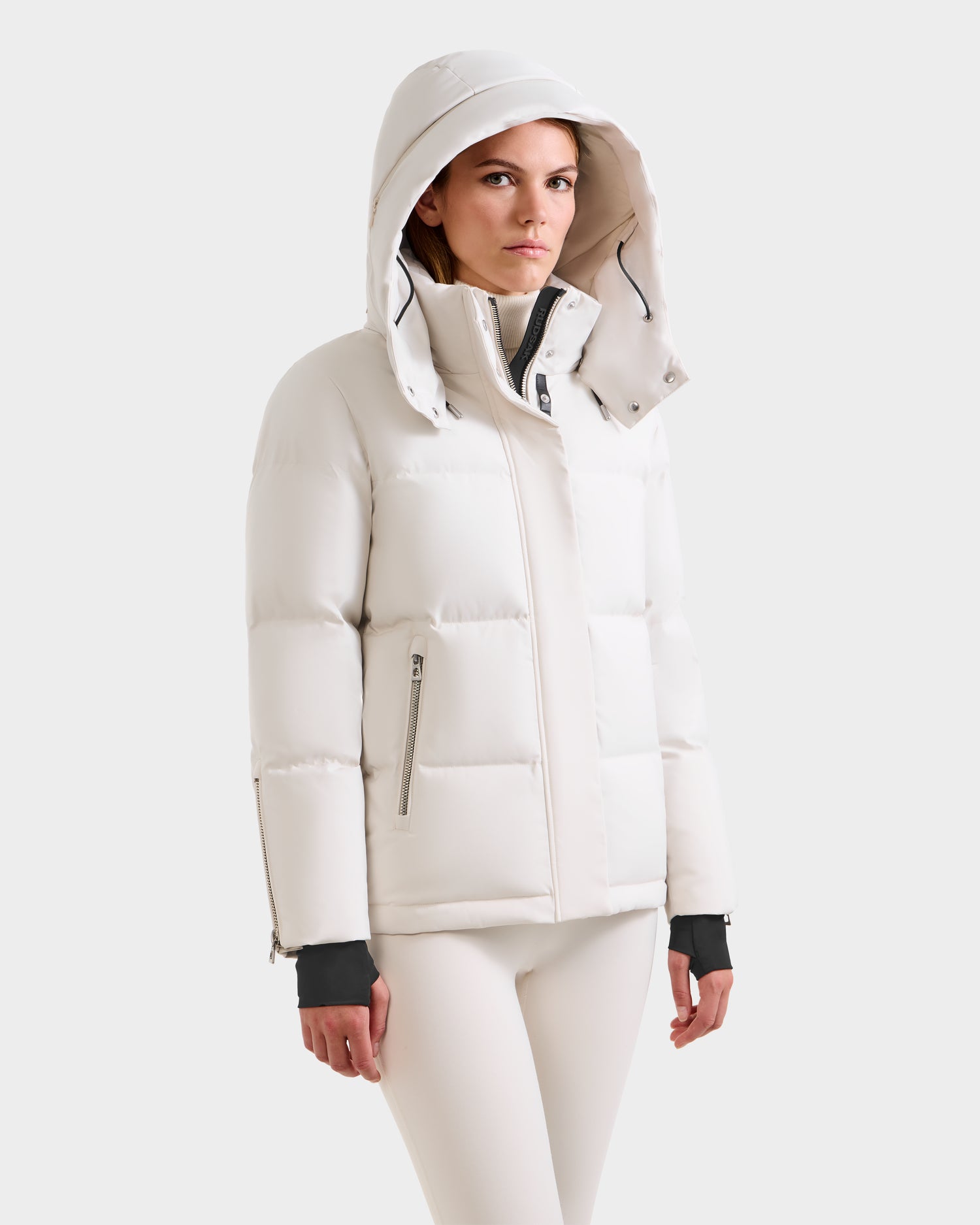 A woman models the Rudsak JOON H - 8125586 Vanilla, a light-colored hooded down jacket with zippered pockets and matching pants—an essential for cold weather—against a plain white background.