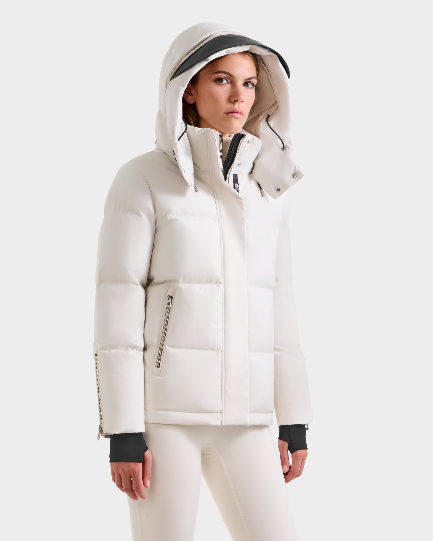 A woman models the Rudsak JOON H - 8125586 Vanilla, a white hooded puffer winter coat with zippered pockets and matching pants, standing against a plain light background and looking slightly to the side with a neutral expression.