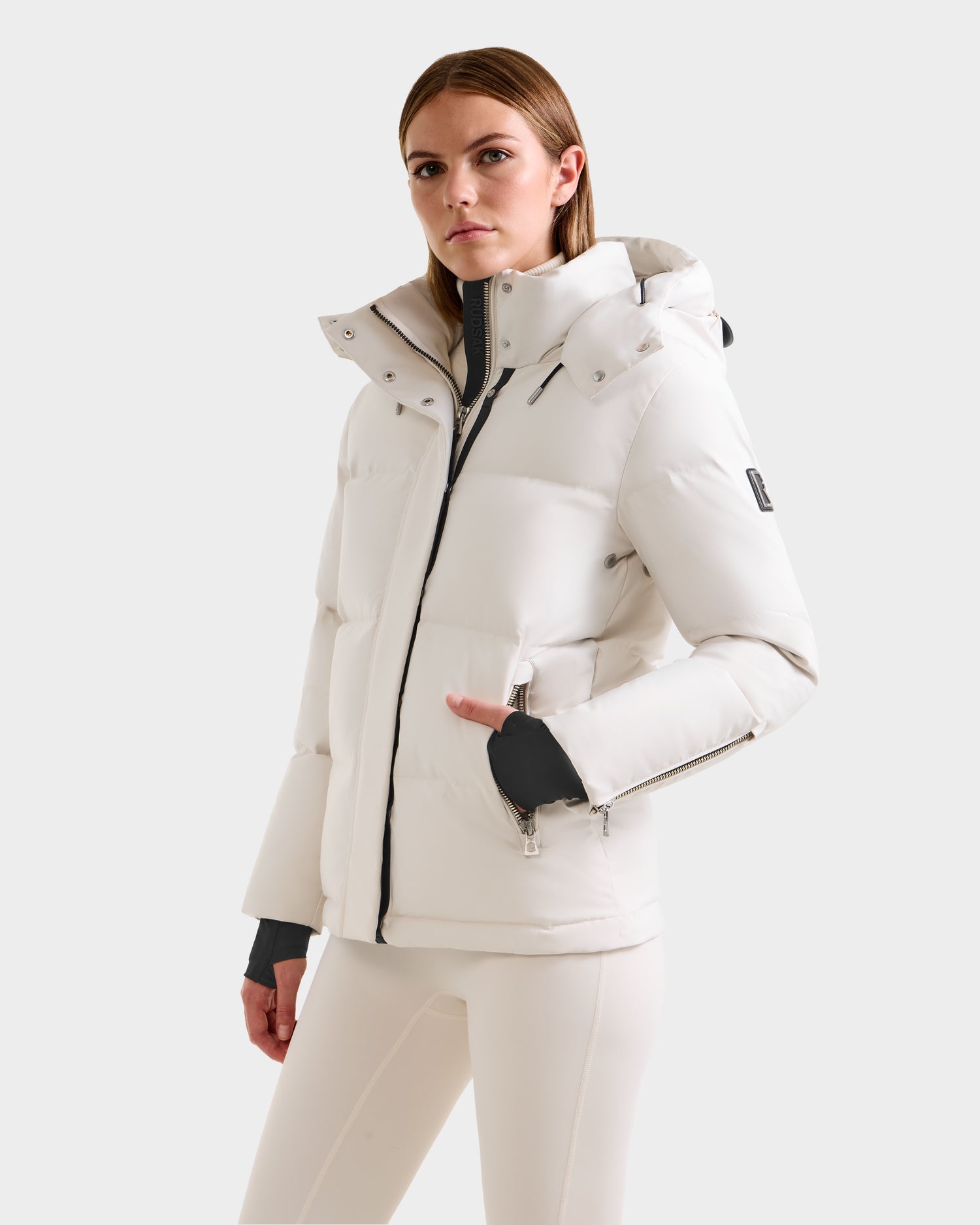 Wearing the Rudsak JOON H - 8125586 Vanilla women’s down jacket with matching pants, a woman stands against a light background, confidently looking at the camera with one hand in her pocket. Lightweight warmth for cold days.
