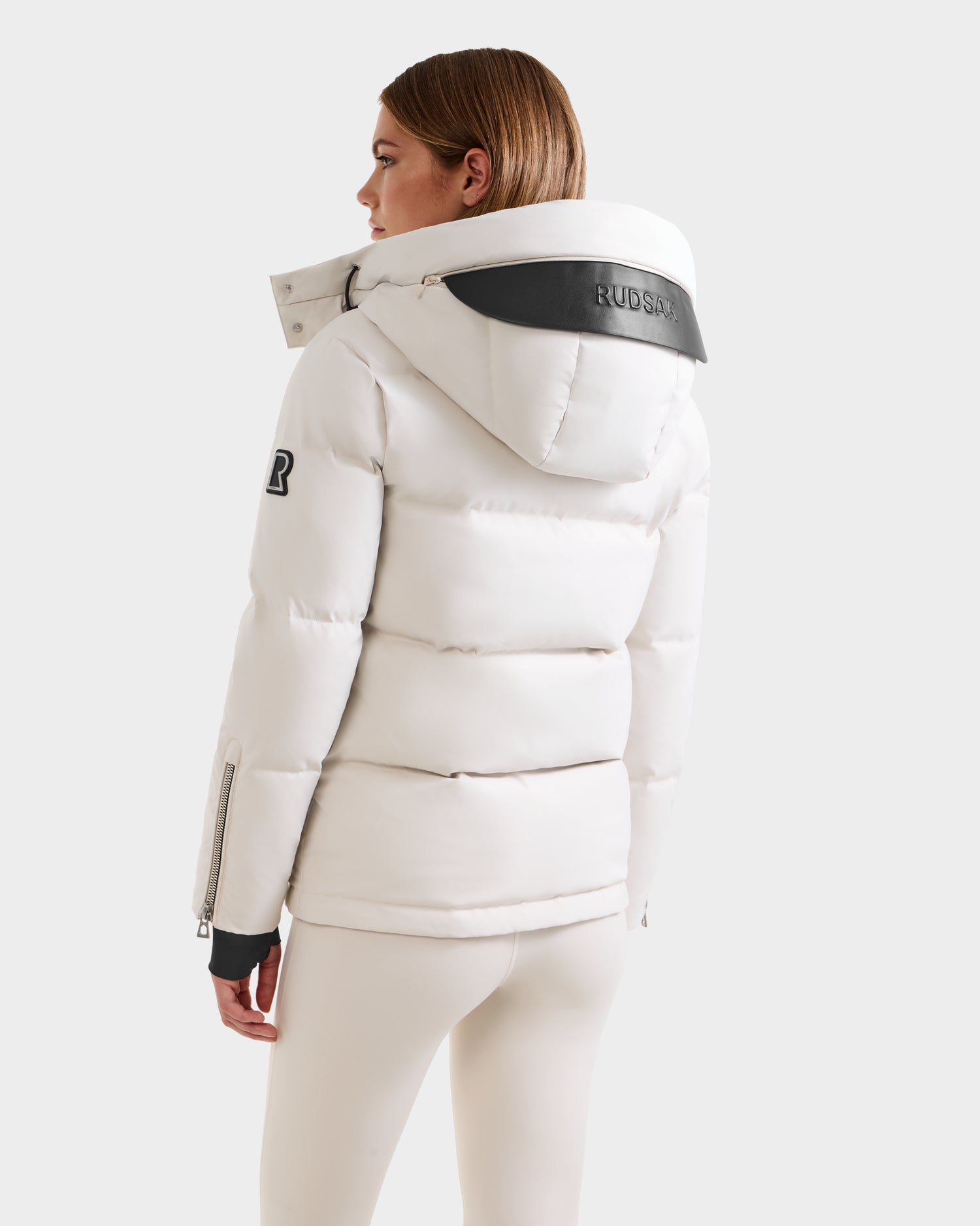 A woman is seen from the back wearing Rudsak’s JOON H - 8125586 Vanilla women’s down jacket with black accents, a logo patch on the sleeve, and a RUDSAK label on the hood, paired with matching white pants.