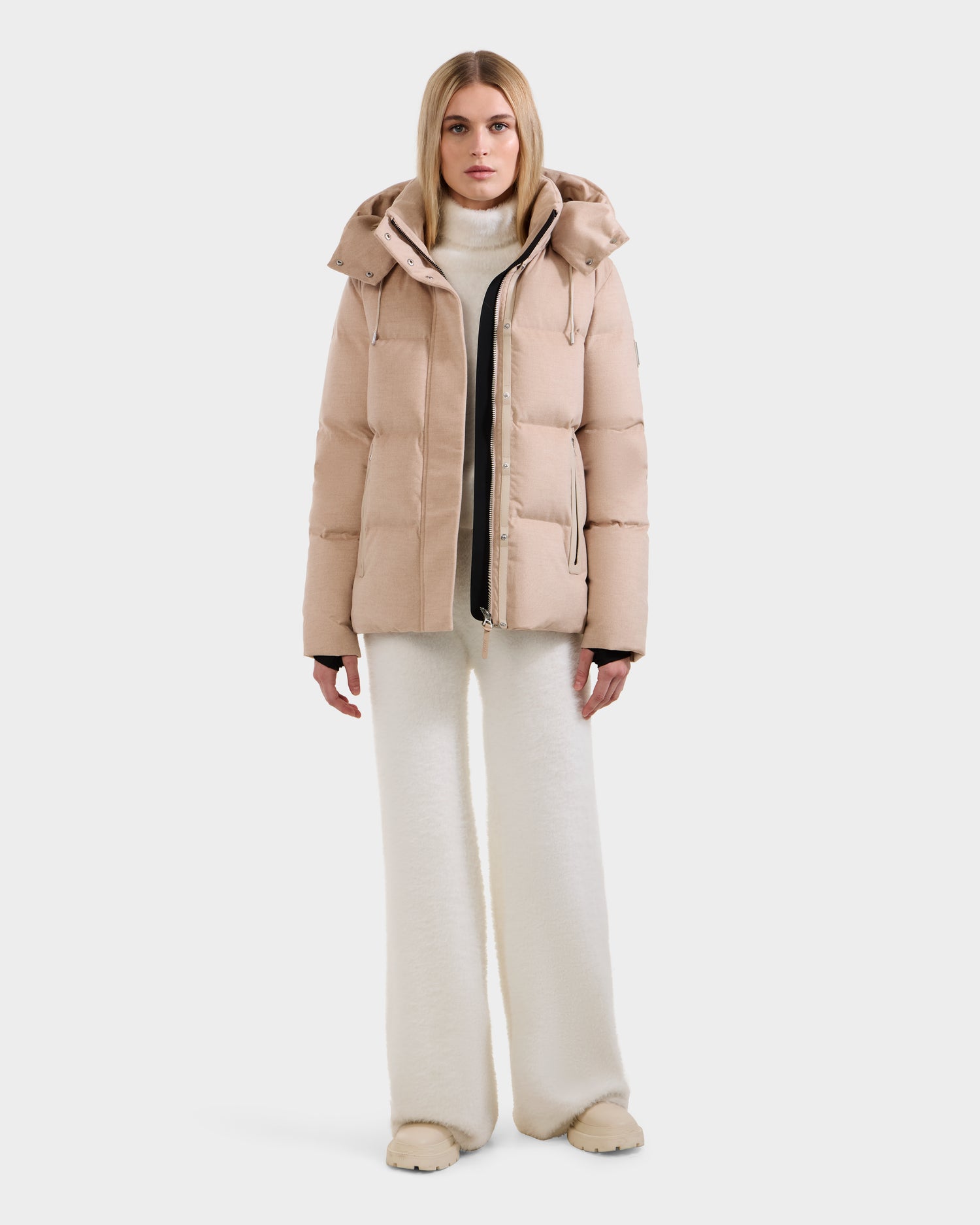 A woman stands facing forward against a plain white background, wearing the Rudsak JOON W - 8125566 premium cold-weather jacket in Heather Beige, paired with white wide-leg pants, a white turtleneck, and beige shoes.