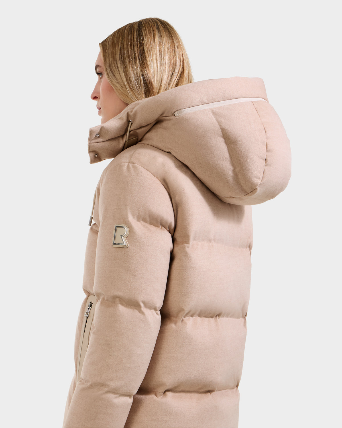 A blonde woman models the Rudsak JOON W - 8125566 Heather Beige wool-blend down puffer with a large hood and zippered pocket, shown in profile against a plain white background.
