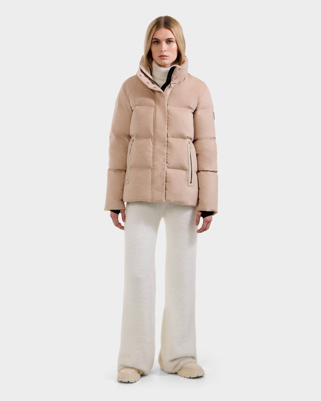A woman stands facing forward against a plain light background, wearing the Rudsak JOON W - 8125566 Heather Beige premium cold-weather jacket, paired with white wide-leg pants and beige shoes.