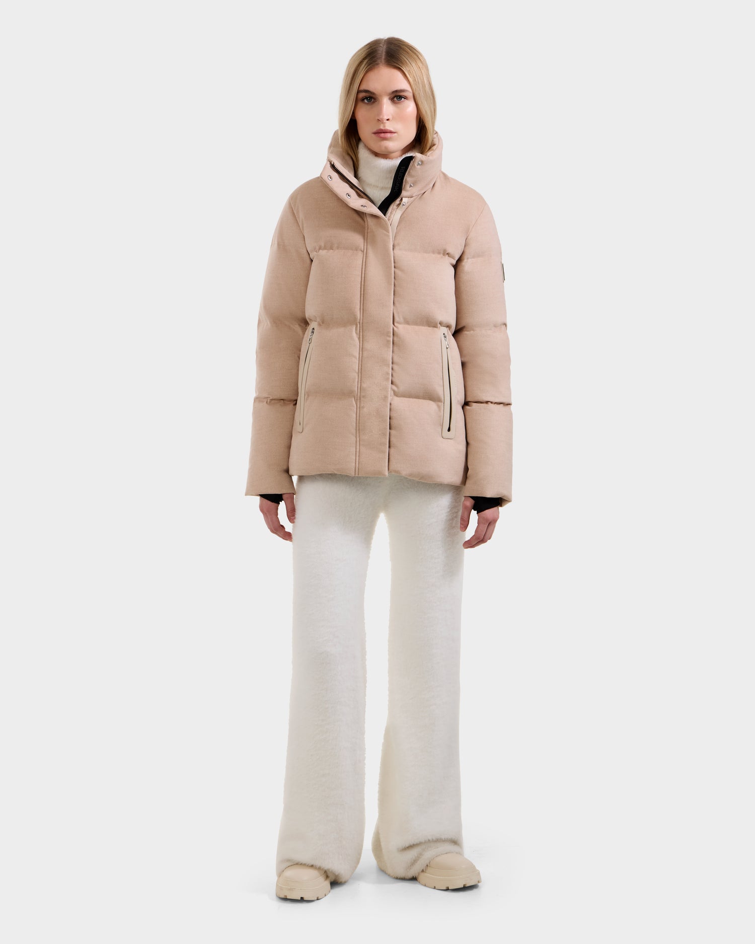 A woman stands facing forward against a plain light background, wearing the Rudsak JOON W - 8125566 Heather Beige premium cold-weather jacket, paired with white wide-leg pants and beige shoes.