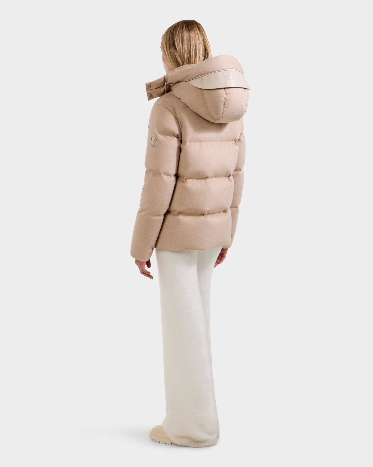 Seen from behind, a blonde woman is wearing the Rudsak JOON W - 8125566 Heather Beige wool-blend down puffer jacket with removable hood and white wide-leg pants against a plain white background.