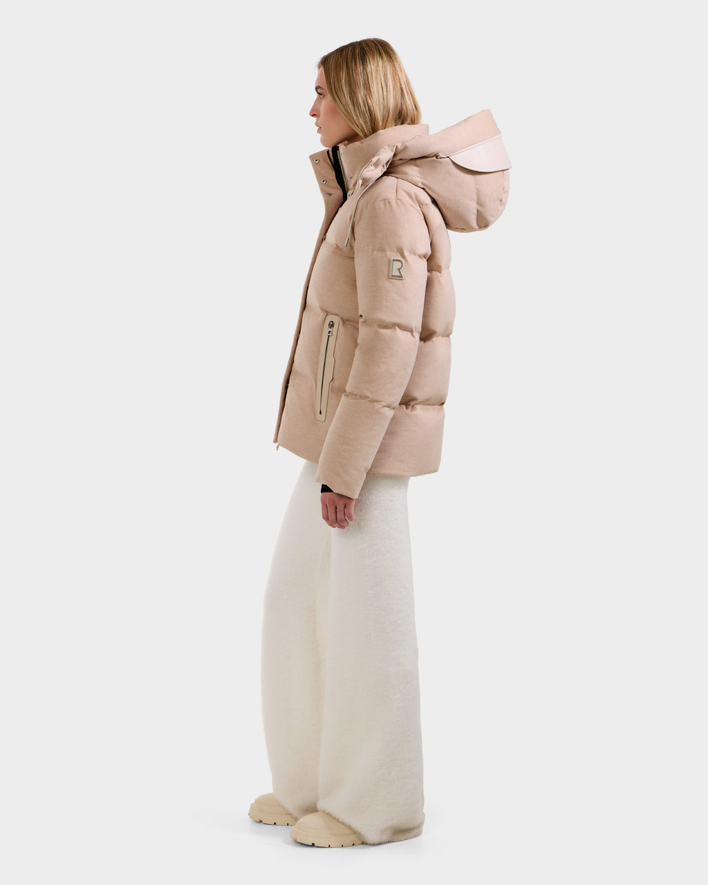 A woman in profile wears the Rudsak JOON W - 8125566 Heather Beige premium cold-weather puffer jacket with a hood, styled with off-white wide-leg pants and beige shoes against a plain white background.