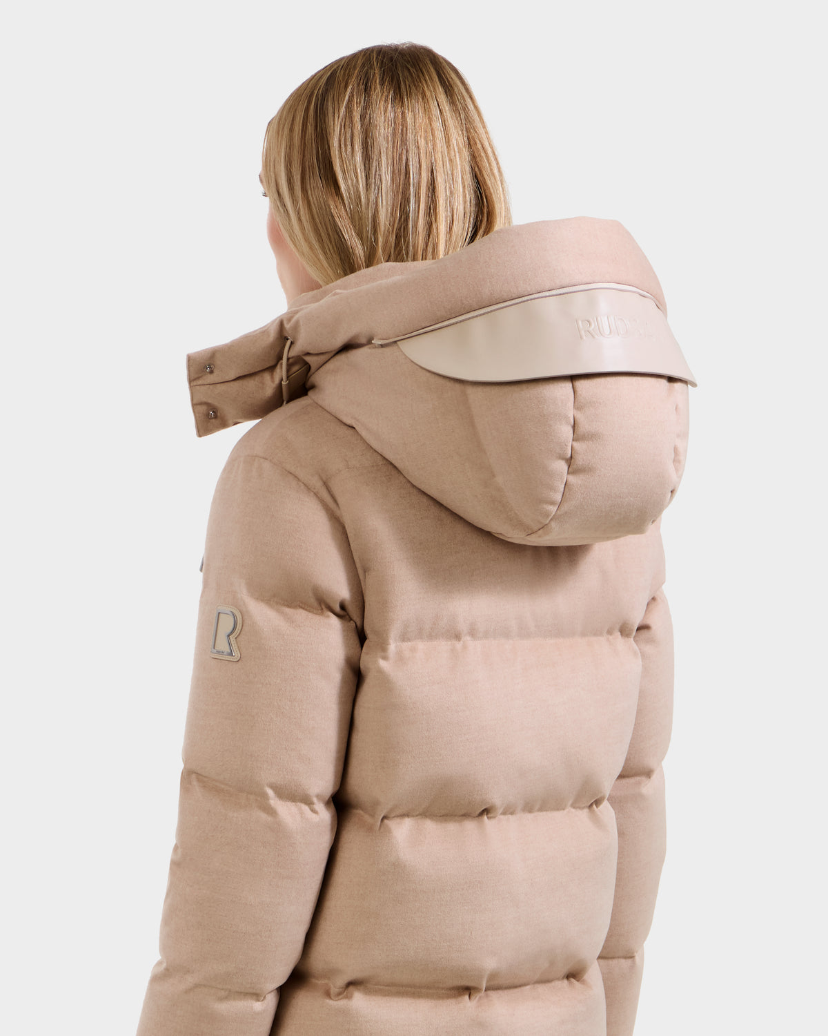 A woman with blonde hair, seen from behind, wears the Rudsak JOON W - 8125566 Heather Beige puffer jacket, featuring a logo patch on the sleeve and a shiny hood accent, set against a light background.