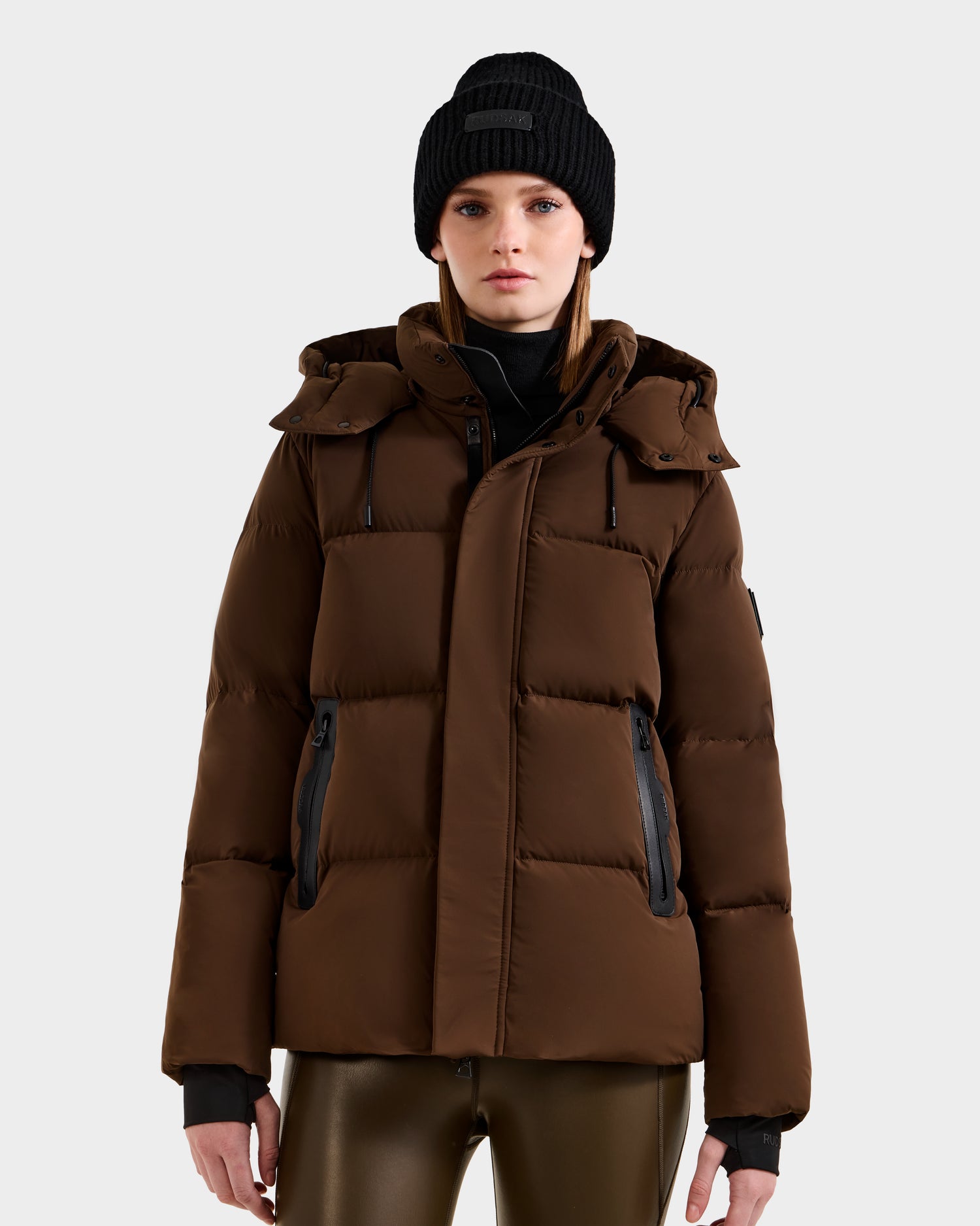 A person stands facing forward against a plain light gray background, wearing the Rudsak JOON - 8124728 Mocha weather-resistant brown down puffer jacket with a hood, a black beanie, and dark shiny pants.
