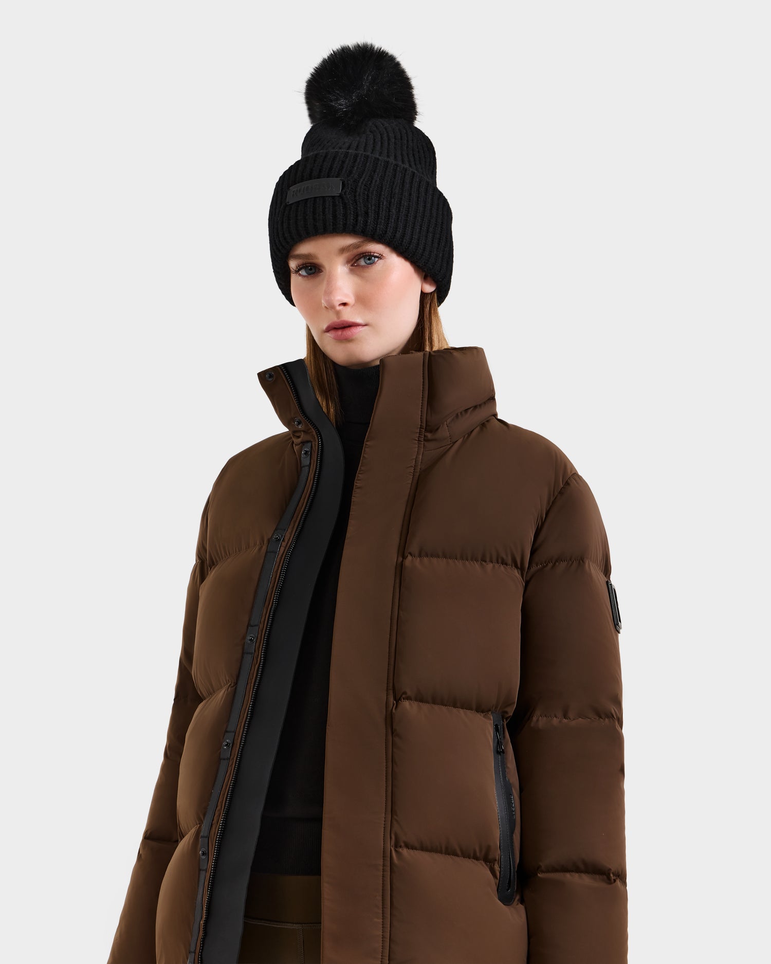 Wearing the Rudsak JOON - 8124728 Mocha brown down puffer jacket and a black pom-pom beanie, this person stands against a plain light background, looking forward with a neutral expression.