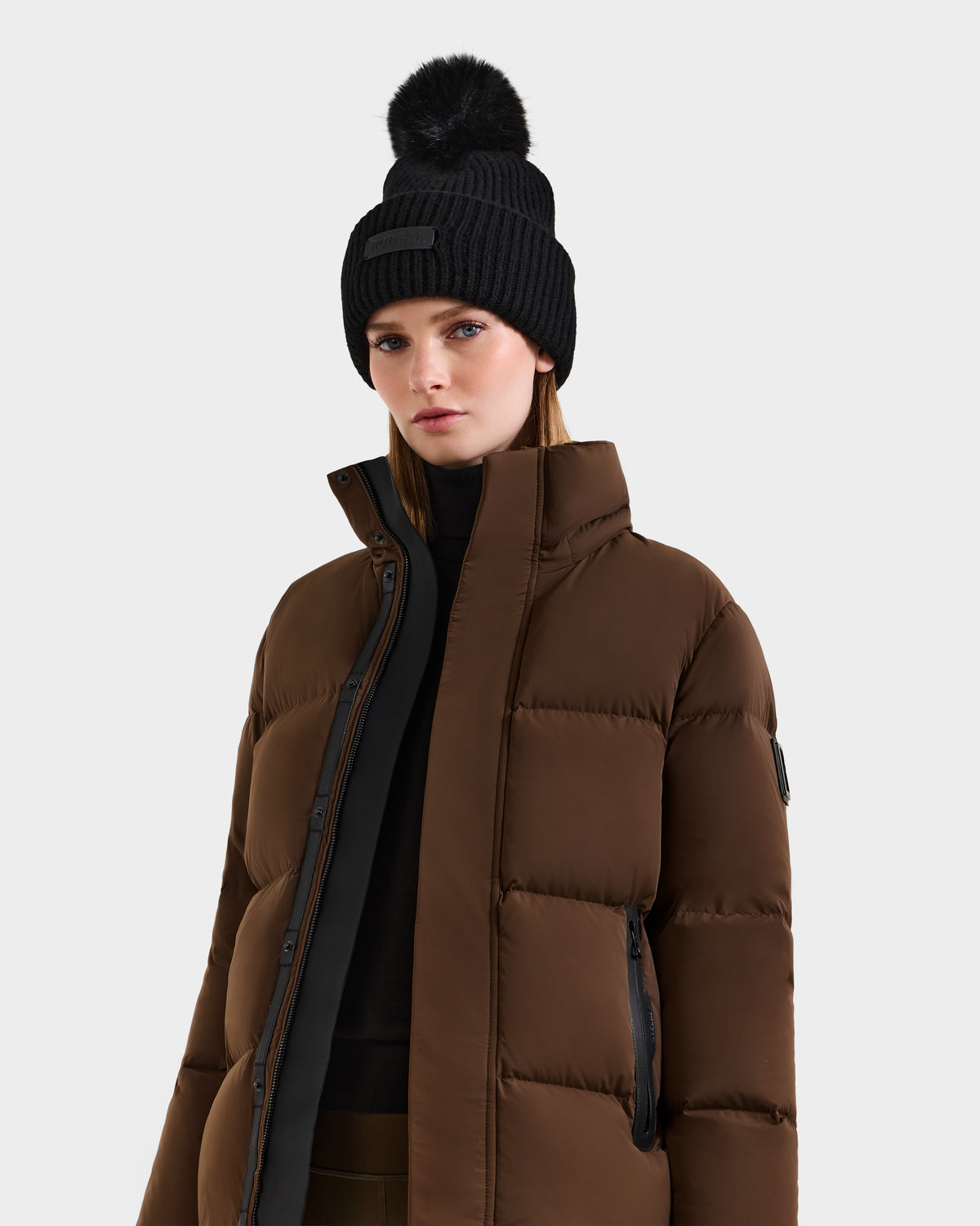 Wearing the Rudsak JOON - 8124728 Mocha brown down puffer jacket and a black pom-pom beanie, this person stands against a plain light background, looking forward with a neutral expression.