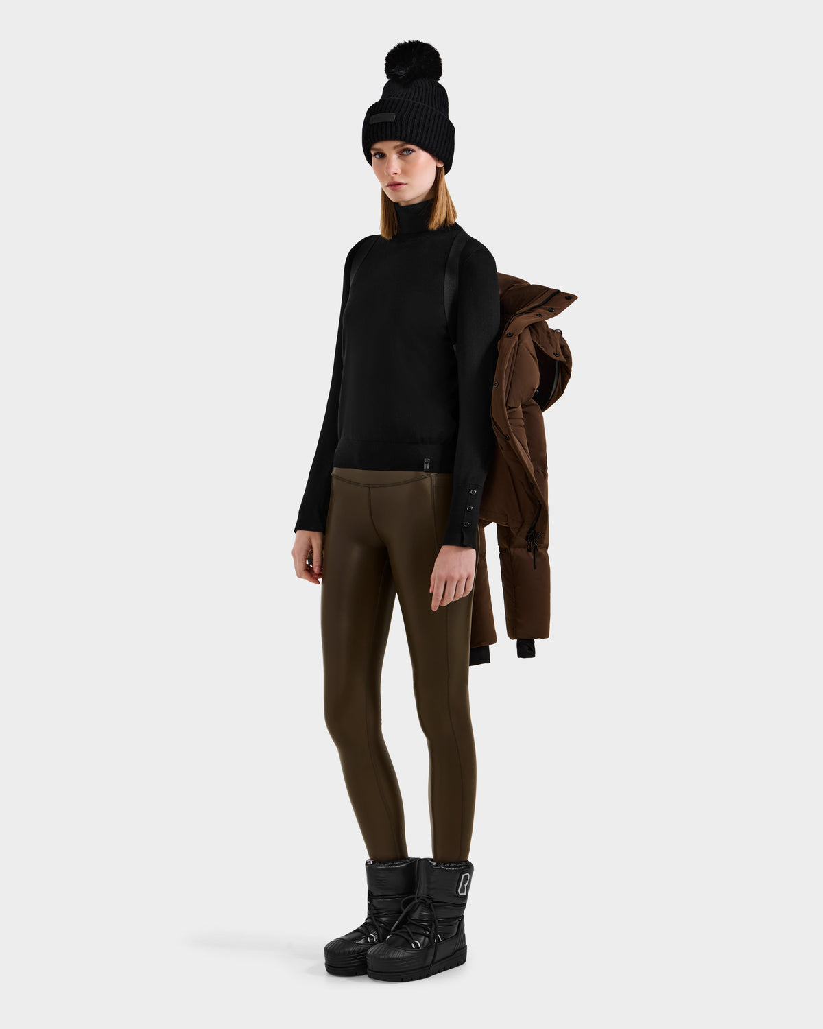 A woman stands against a plain background wearing the Rudsak JOON - 8124728 Mocha weather-resistant coat draped over her shoulder, paired with a black beanie, black sweater, olive leggings, and black winter boots.