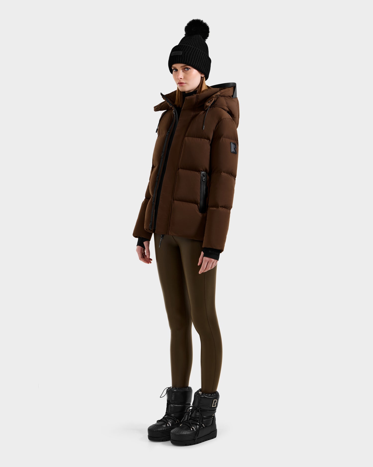 A person stands against a plain background, wearing the Rudsak JOON - 8124728 Mocha brown down puffer jacket, olive green leggings, black winter boots, and a black knit hat with a pom-pom.