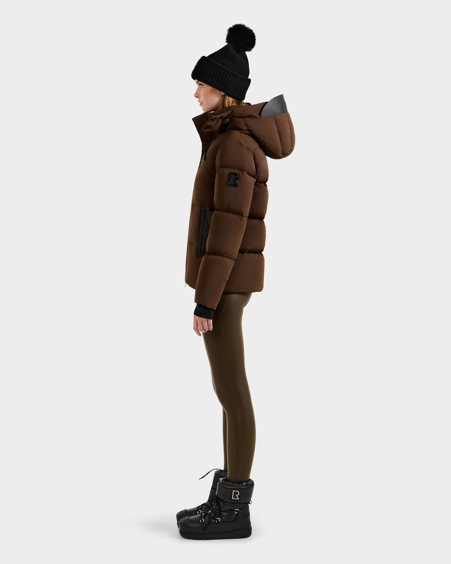 A person stands in profile against a plain light background wearing the Rudsak JOON - 8124728 Mocha down puffer jacket with a hood, olive leggings, black winter boots, black gloves, and a black knit pom-pom beanie.