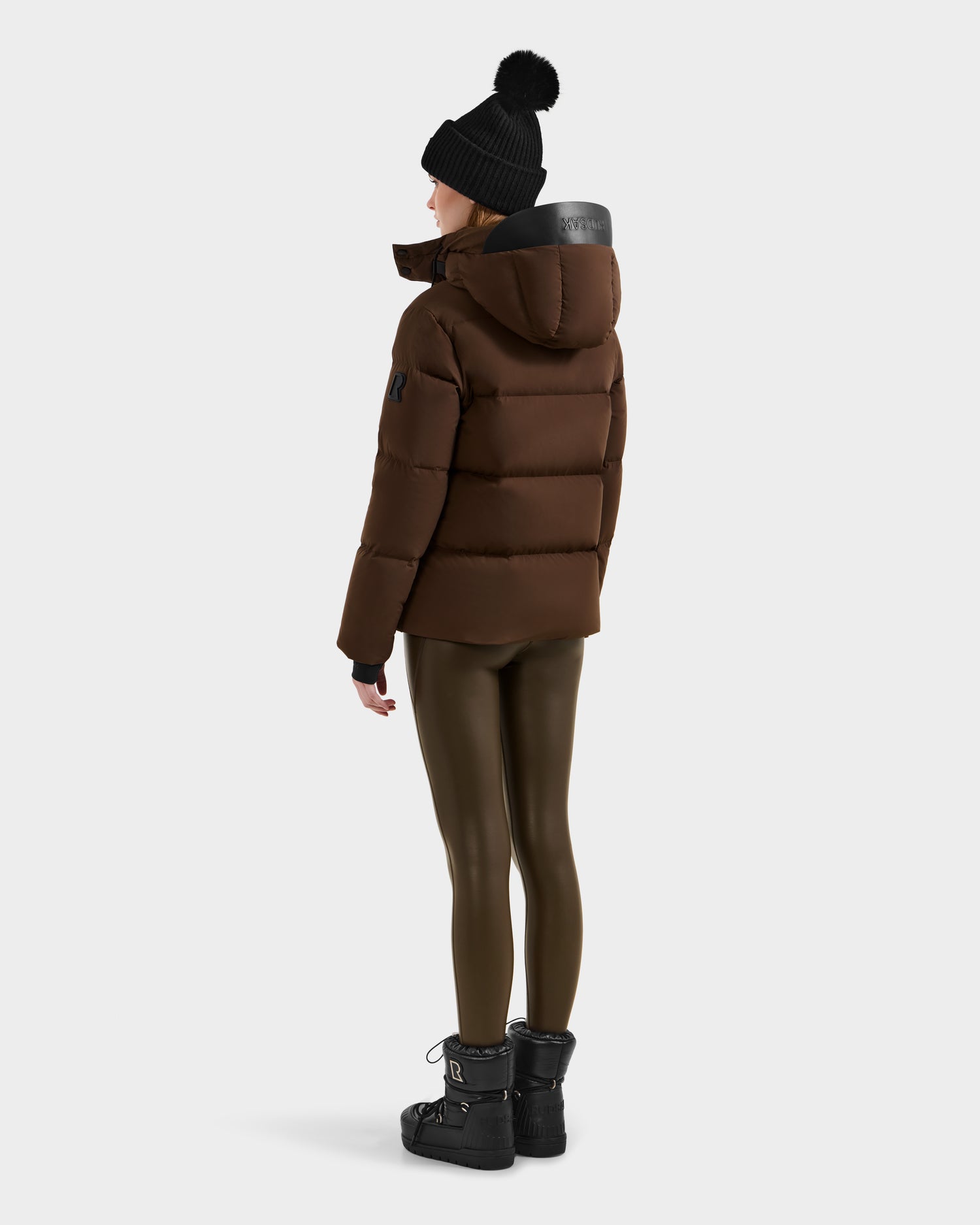 A person stands facing away, wearing the Rudsak JOON - 8124728 Mocha down puffer jacket with hood, on a plain light background.