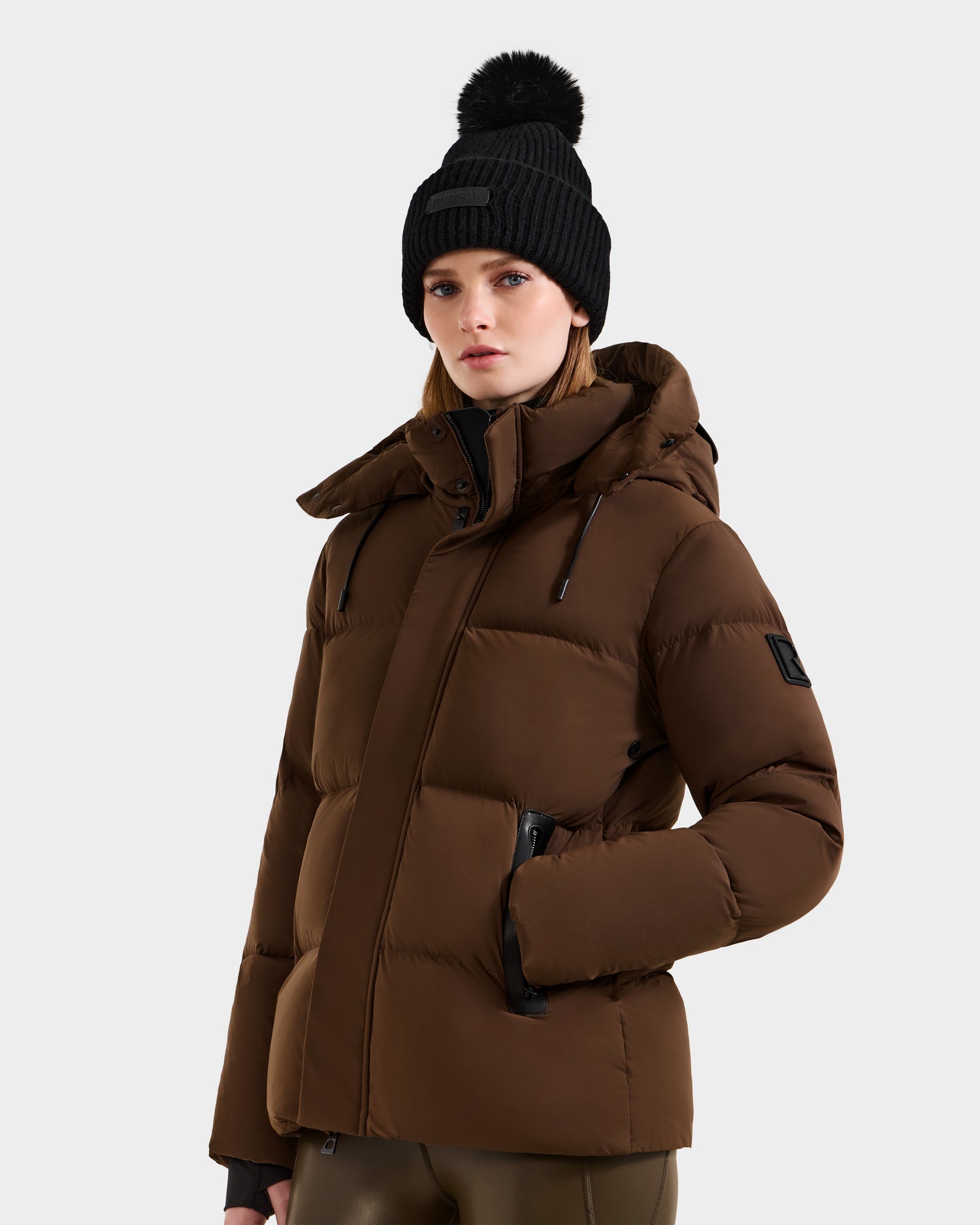 A woman wearing the Rudsak JOON - 8124728 Mocha down puffer jacket and a black knit beanie with a pom-pom stands against a plain light background, facing forward with a neutral expression.