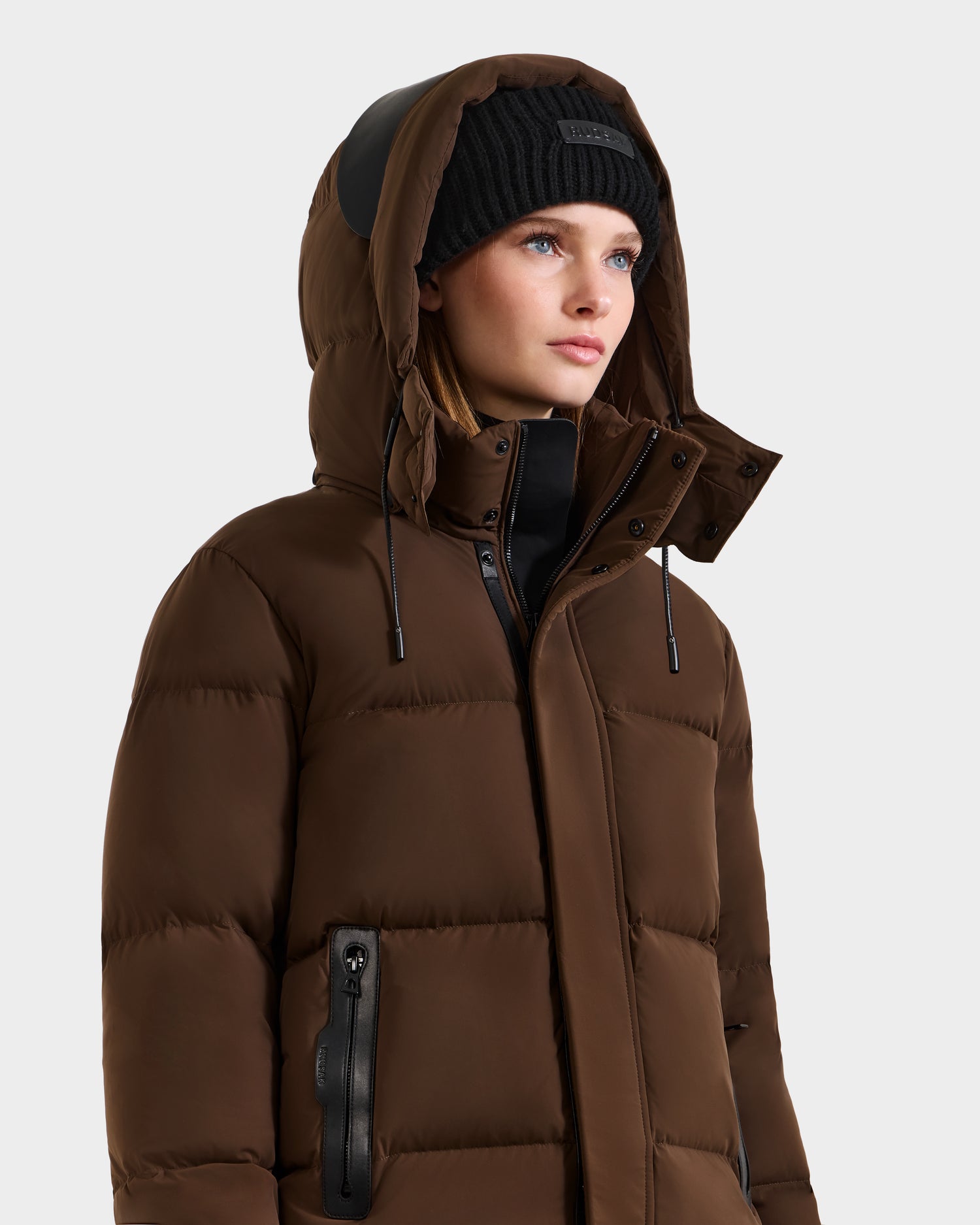 A woman wears the Rudsak JOON - 8124728 Mocha weather-resistant down puffer jacket with a hood and a black knit beanie, gazing upward against a plain light background.