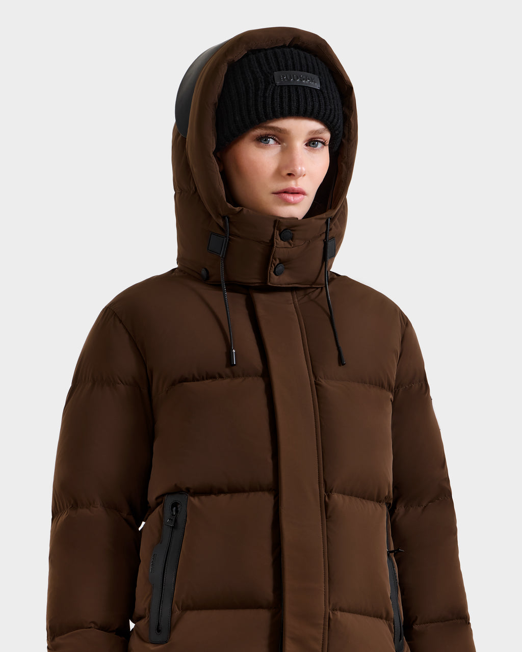 A woman in the Rudsak JOON - 8124728 Mocha weather-resistant down puffer jacket with a hood and a black knit beanie stands against a plain light gray background.