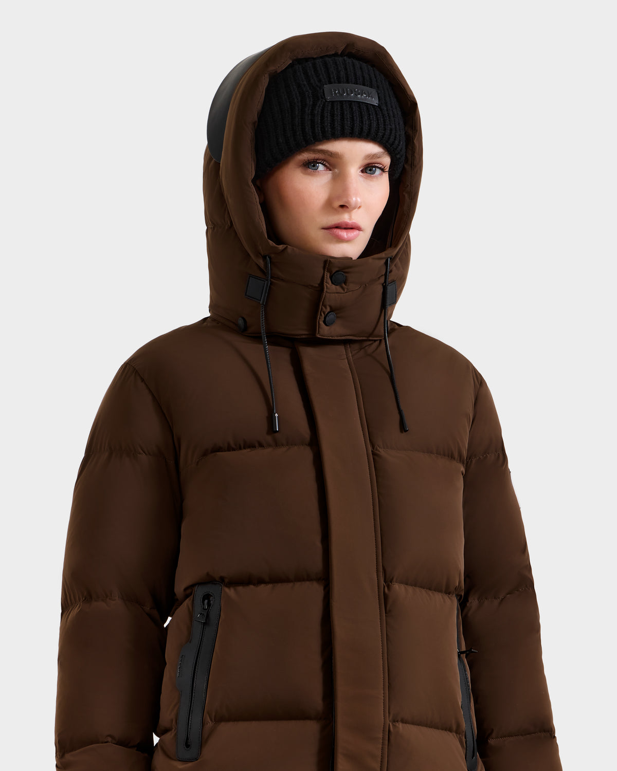 A woman in the Rudsak JOON - 8124728 Mocha weather-resistant down puffer jacket with a hood and a black knit beanie stands against a plain light gray background.