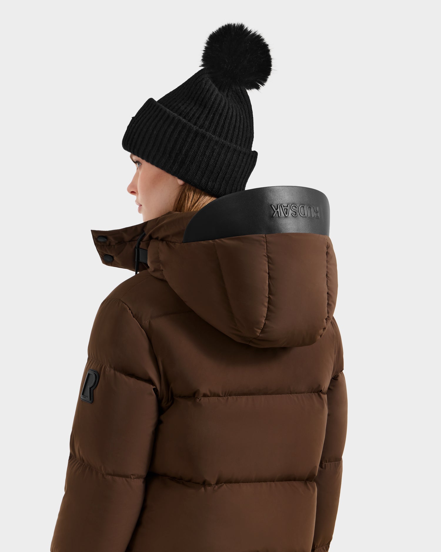 A person wearing the Rudsak JOON - 8124728 Mocha down puffer jacket with a hood and a black knit beanie with pom-pom, facing away from the camera against a plain background.