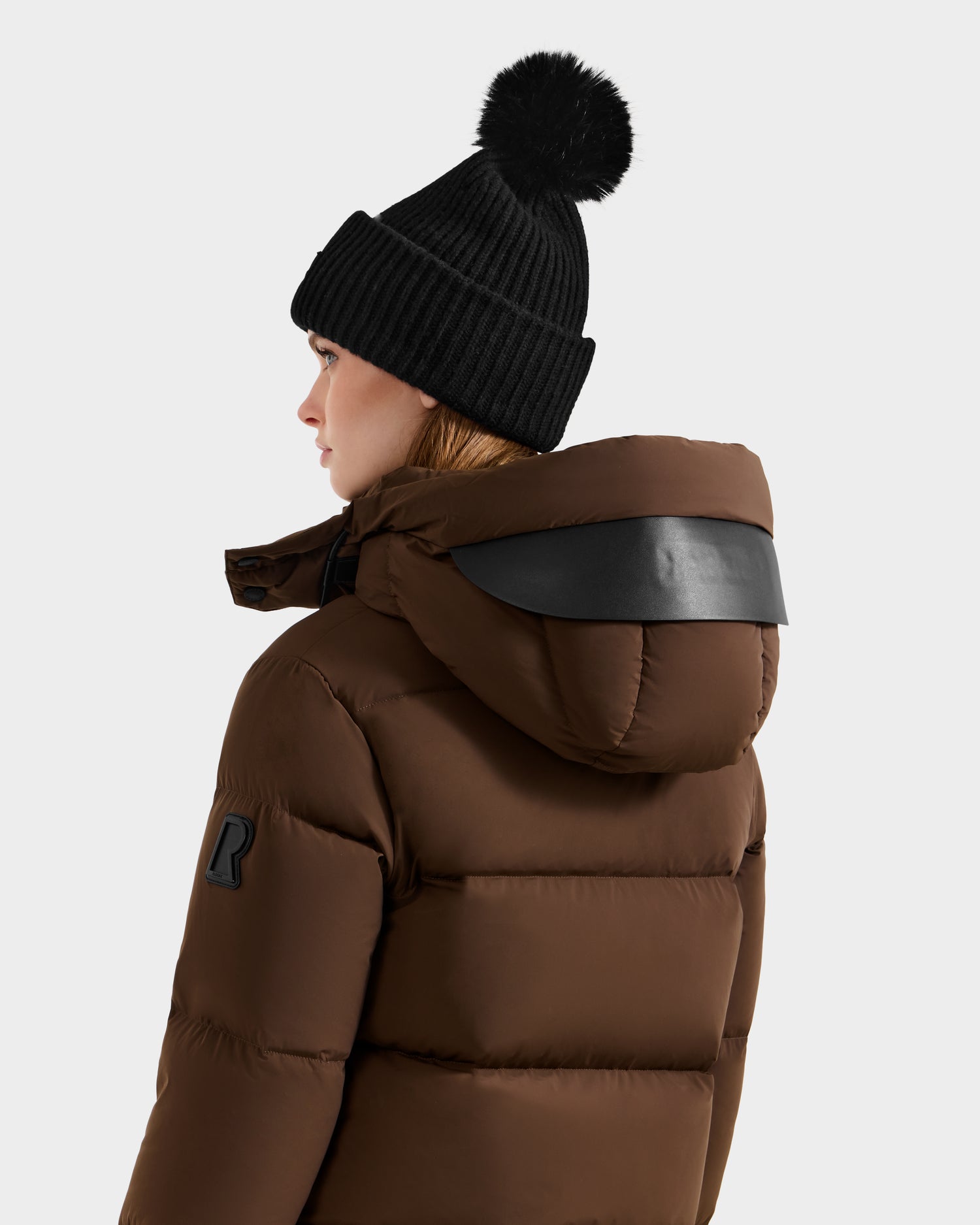 A person in a black knit pom-pom beanie and the Rudsak JOON - 8124728 Mocha RDS-certified down hooded puffer jacket, shown from the side and back against a plain light background.