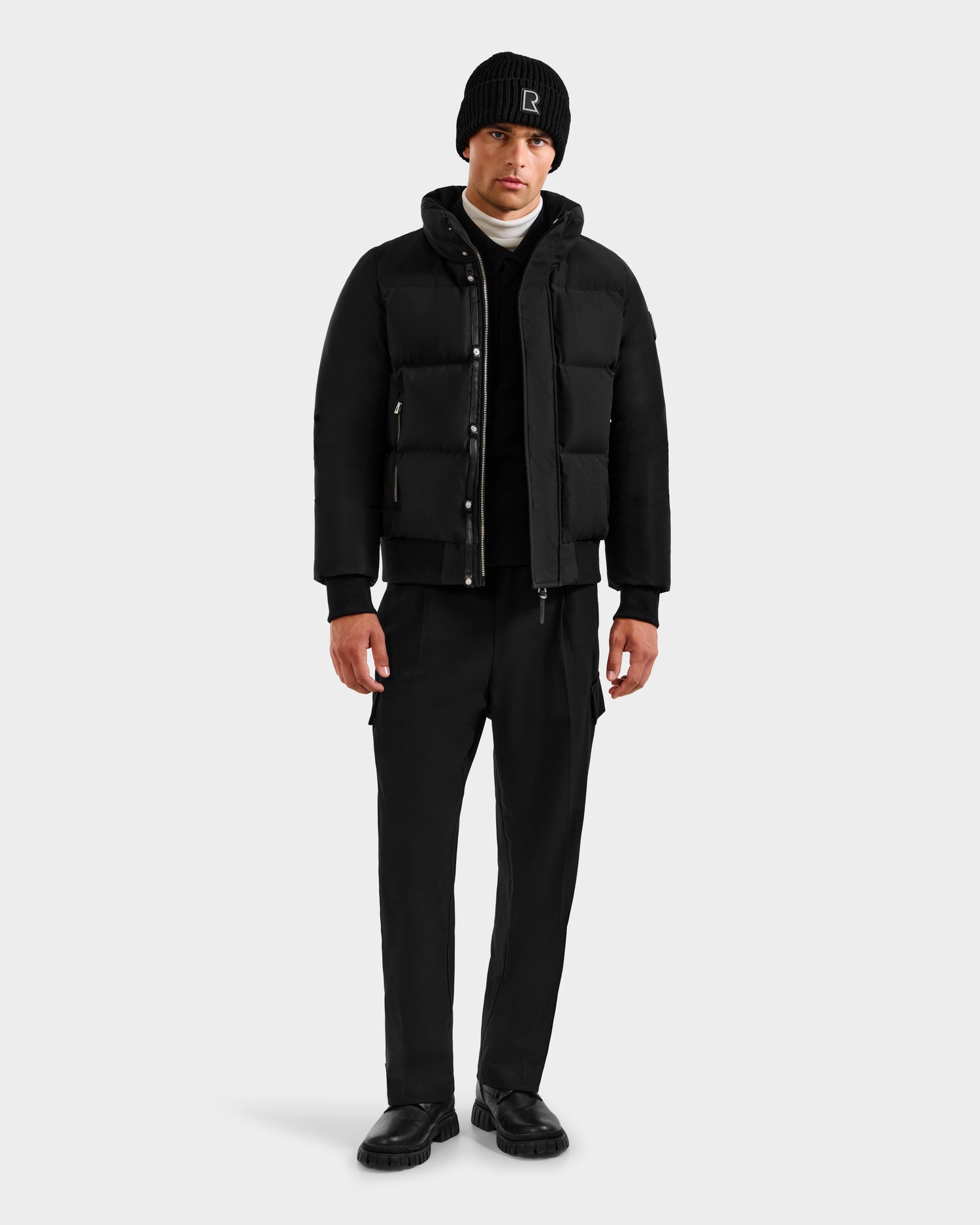 A man stands facing forward wearing the Rudsak JORDY H - 6125689 Black RDS-certified down puffer jacket with a black beanie, white turtleneck, black pants, and black shoes against a plain light gray background.