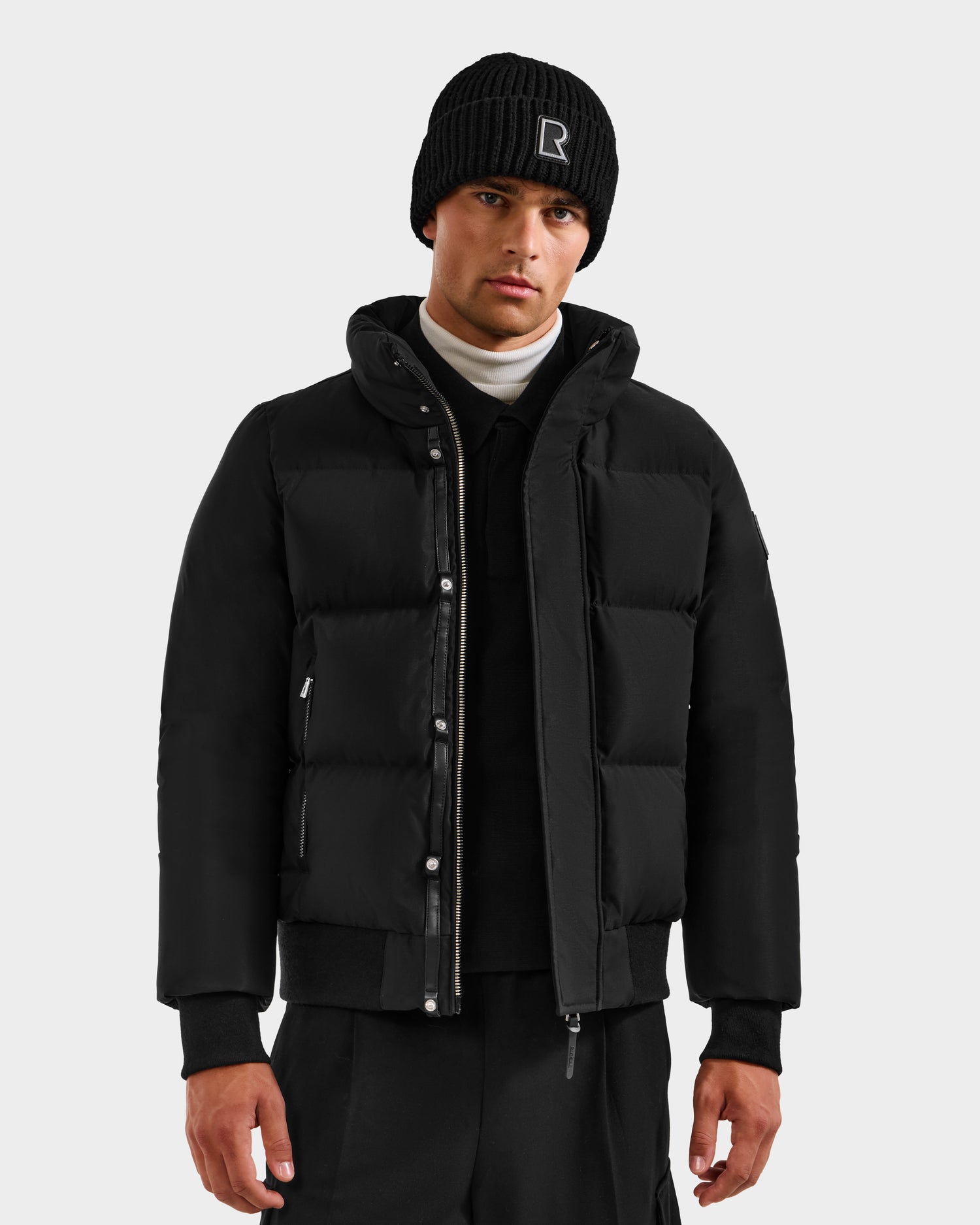 A man wearing the Rudsak JORDY H - 6125689 Black down bomber, a white turtleneck, and black pants stands facing forward against a plain light gray background.