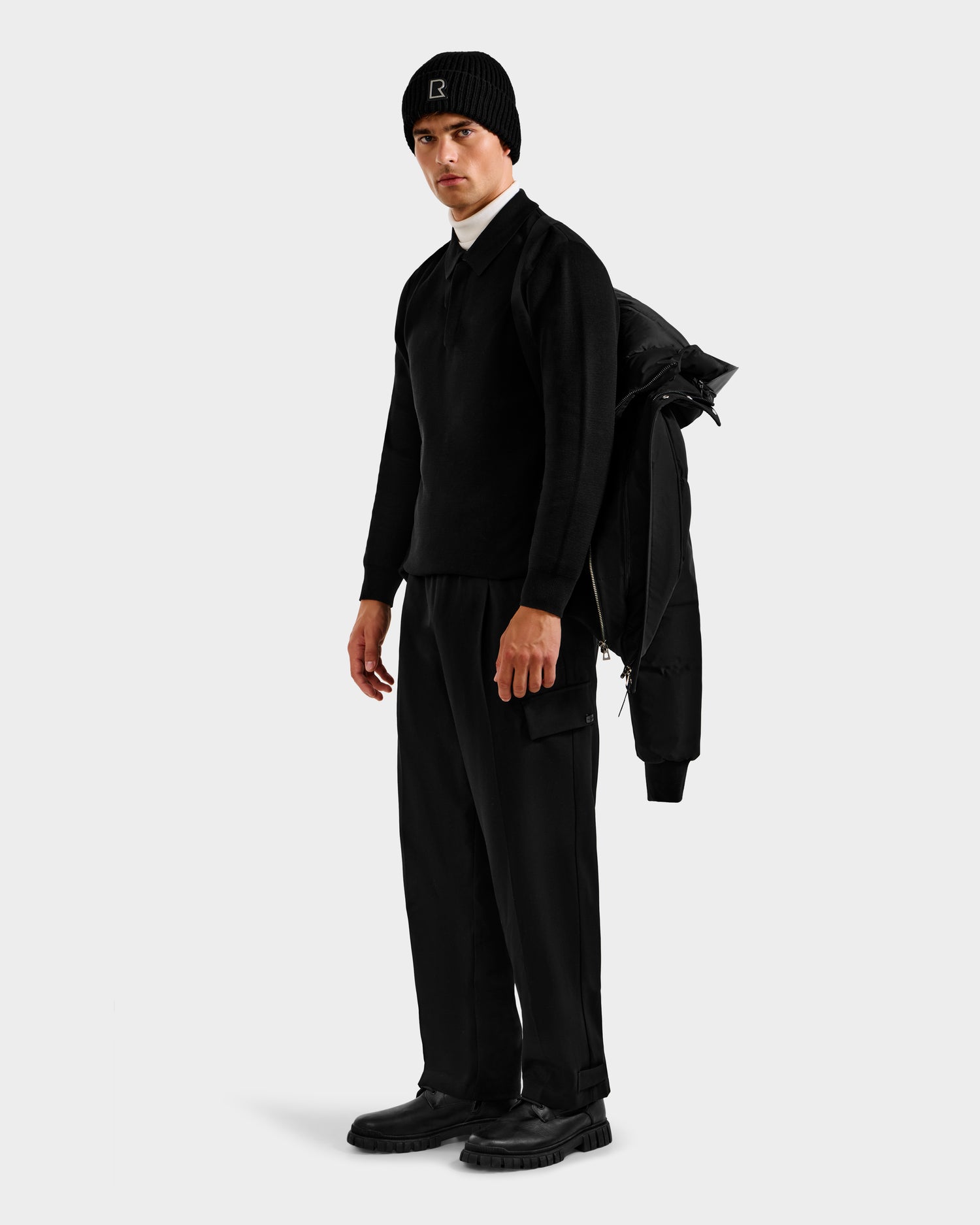 A man holds the Rudsak JORDY H - 6125689 Black quilted bomber jacket over his shoulder, wearing all black, against a plain white background.