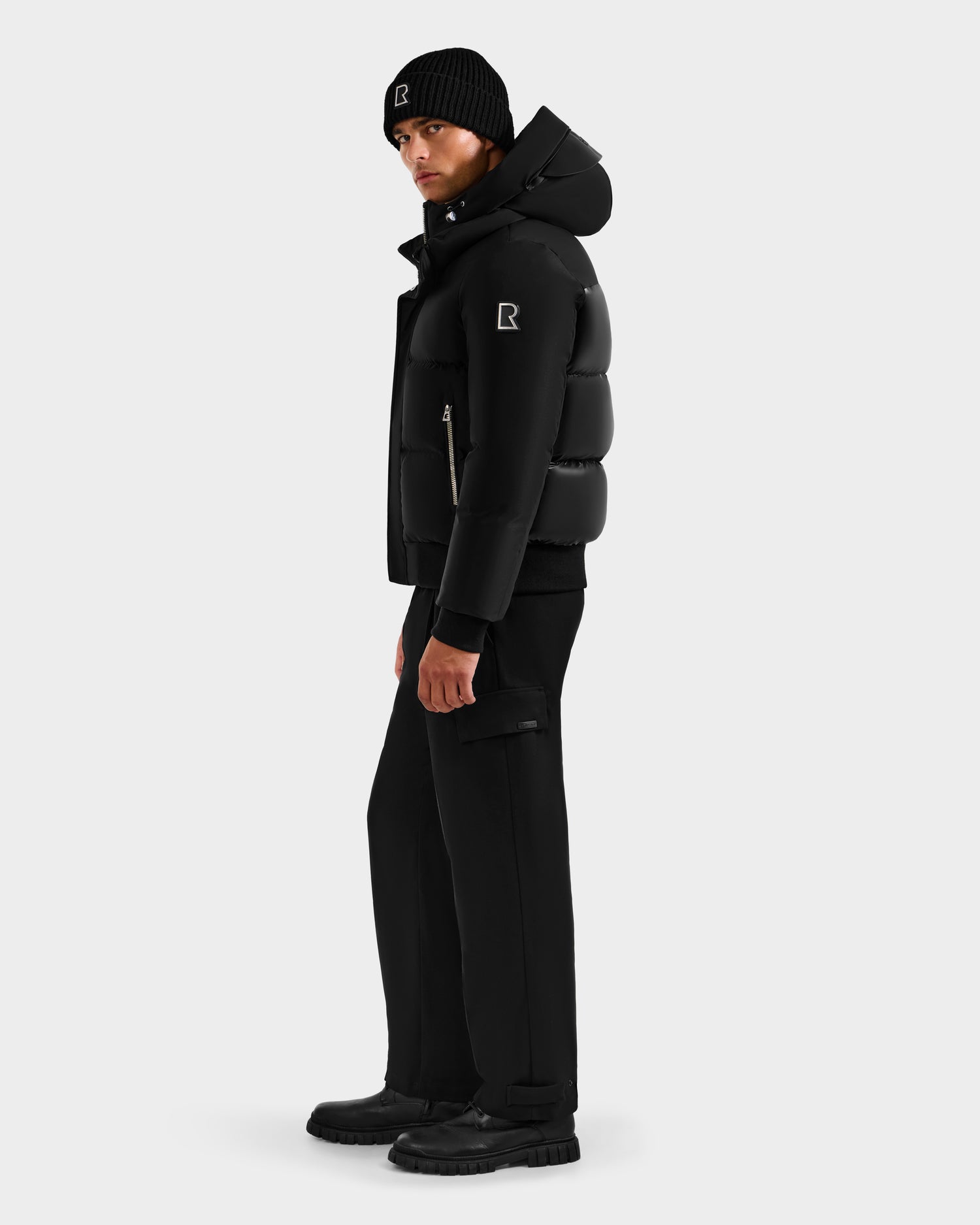 A man stands sideways in an all-black winter outfit by Rudsak, featuring the JORDY H - 6125689 Black down bomber jacket with an R logo on the sleeve and hat. He wears black pants and boots against a plain light gray background.