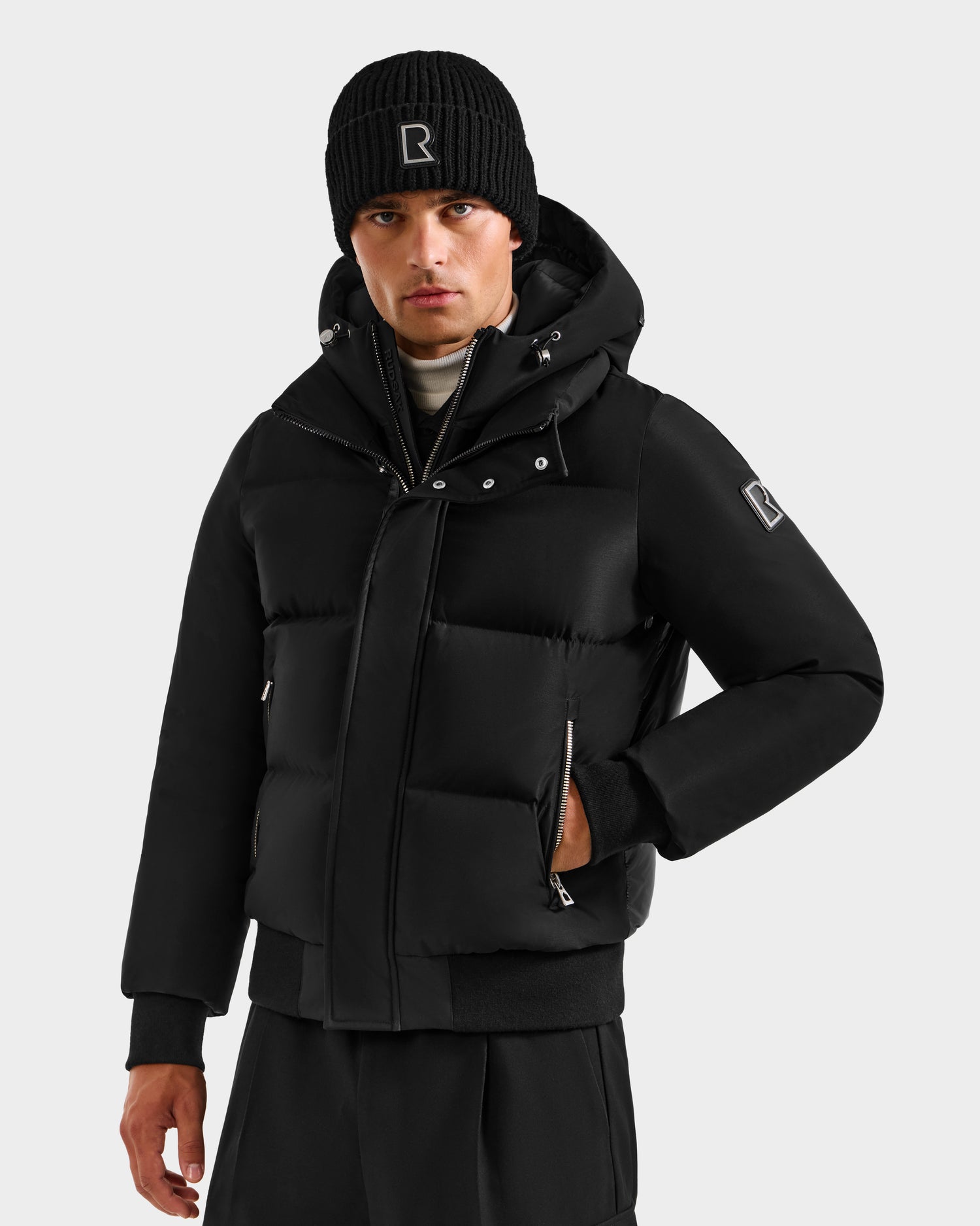 A man wears the Rudsak JORDY H - 6125689 Black quilted down bomber with hood and a black beanie, both featuring the R logo. He stands facing forward, one hand in his coat pocket, against a plain background.