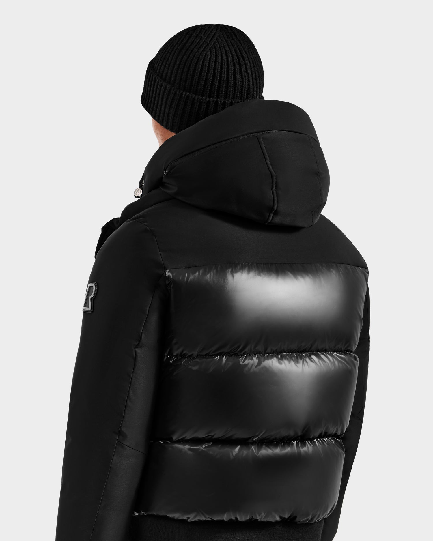 Seen from behind against a plain light background, a person wears a black knit beanie and the Rudsak JORDY H - 6125689 Black men’s quilted down bomber with hood.
