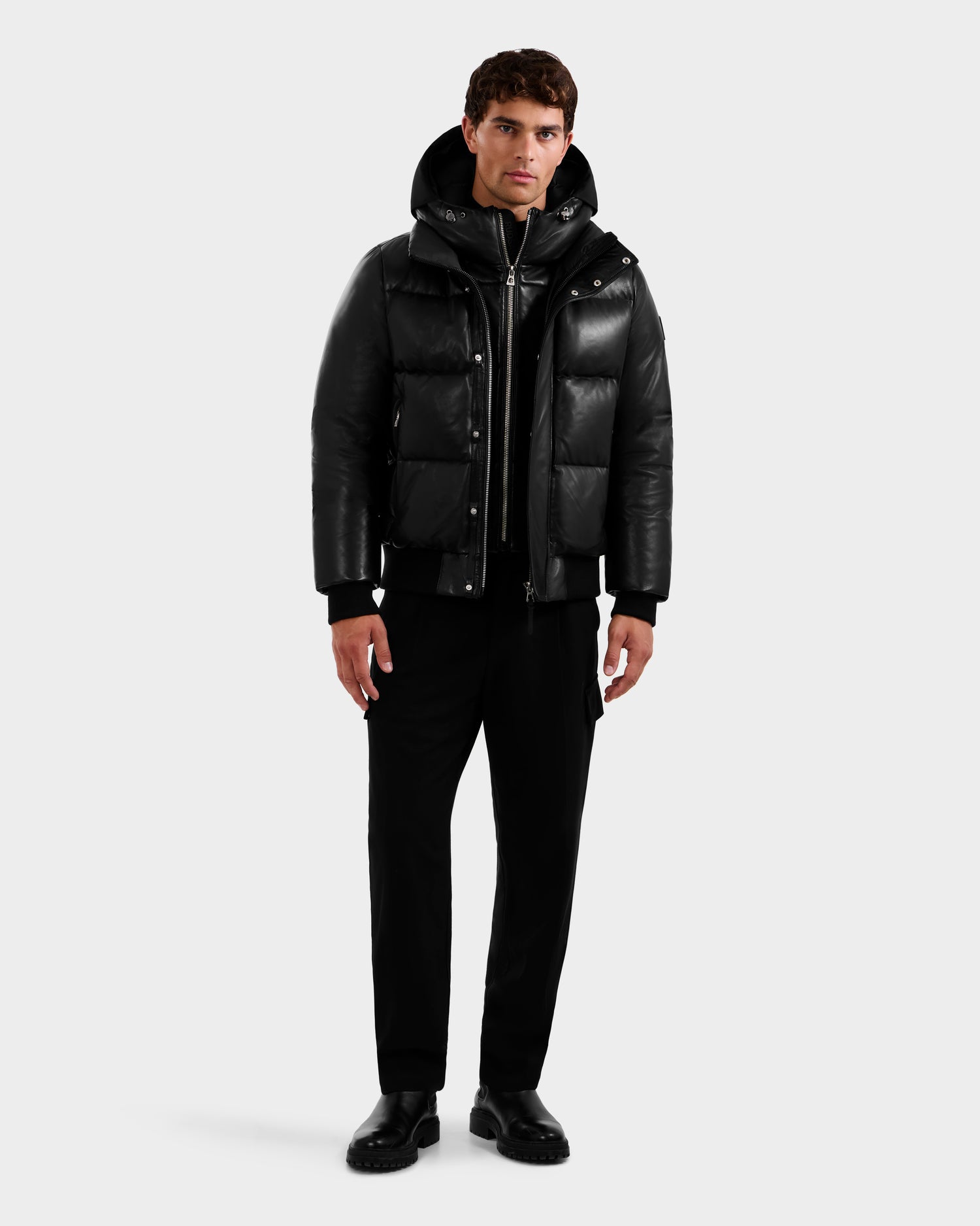 Wearing the Rudsak JORDY X - 6125694 Black unisex puffer jacket with a hood, a person stands facing forward against a plain light background, paired with black zip-up top, pants, and shoes.