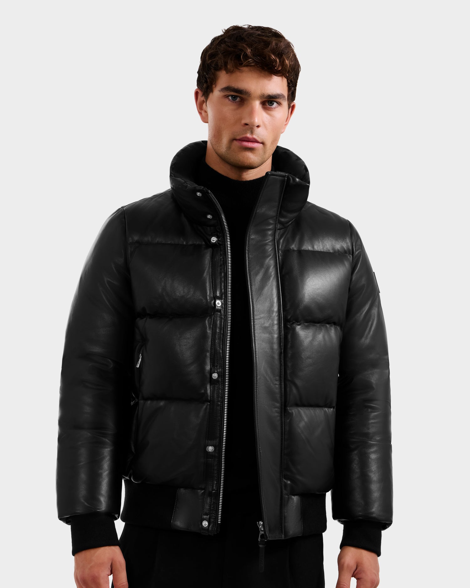 A young man stands against a light gray background, wearing the Rudsak JORDY X - 6125694 Black unisex jacket over a black turtleneck, looking directly at the camera with a neutral expression.