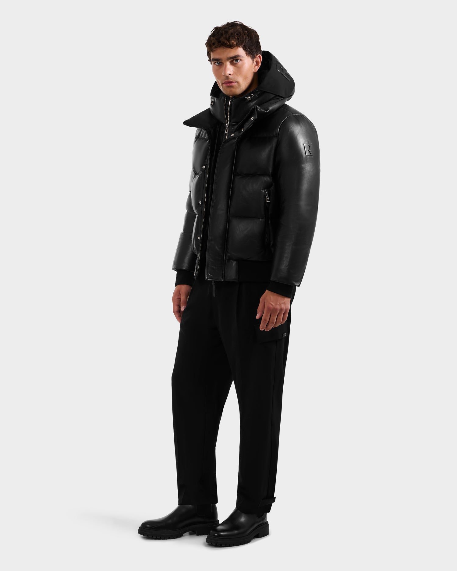 A man wears the Rudsak JORDY X - 6125694 Black quilted bomber with hood, black pants, and black boots, standing against a plain light gray background and looking slightly to the side.