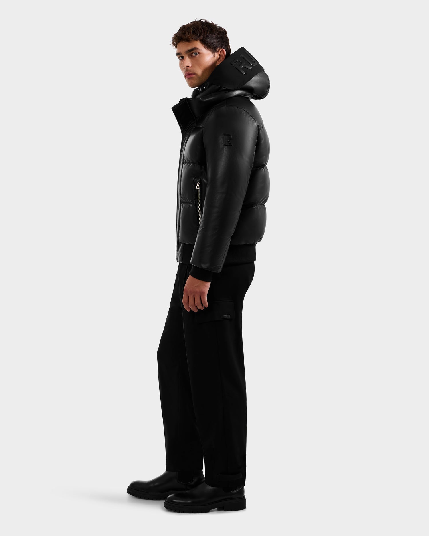 A man poses against a plain light gray background wearing the Rudsak JORDY X - 6125694 Black quilted bomber jacket with a hood, paired with black pants and black shoes.
