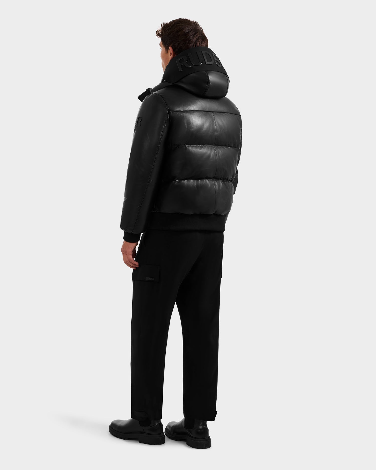 A man faces away wearing the Rudsak JORDY X - 6125694 Black quilted bomber with a hood, paired with black pants and shoes, set against a plain light gray background.