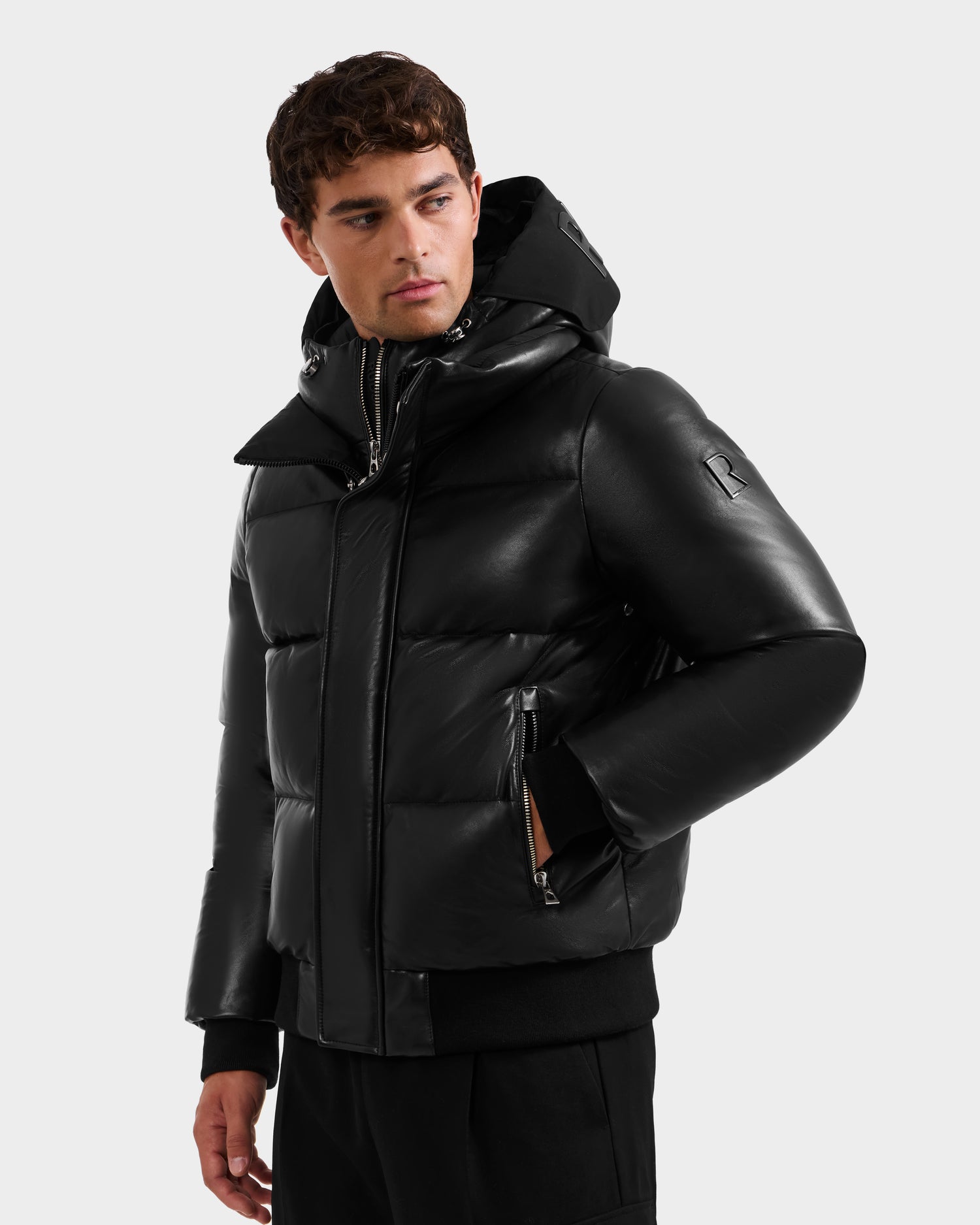 A man in the Rudsak JORDY X - 6125694 Black shiny unisex quilted bomber with a hood and zippered pockets, paired with black pants, stands against a plain light gray background looking slightly to his left.