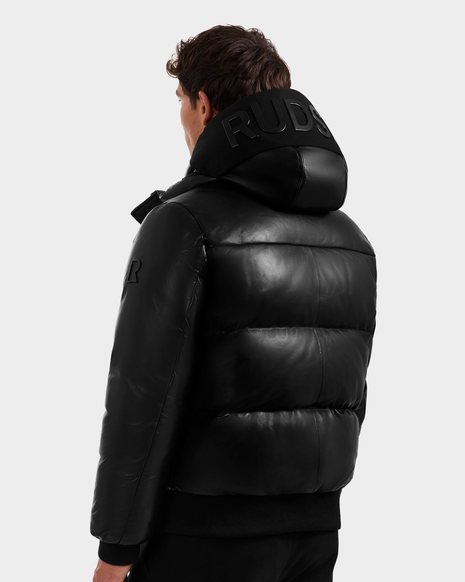 A man stands facing away, showcasing the back and side of the unisex JORDY X - 6125694 Black quilted bomber by Rudsak, featuring an embossed hood, set against a plain light gray background.
