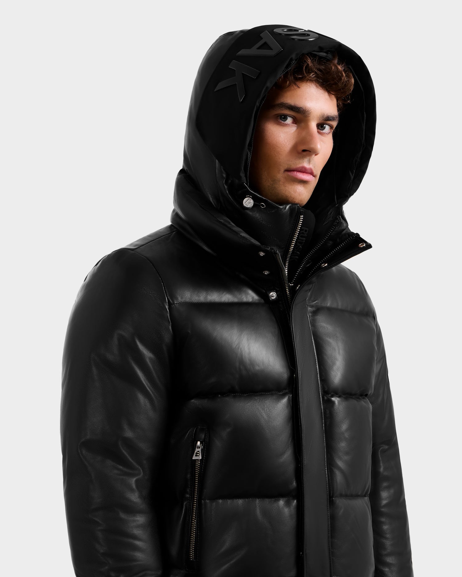 A young man with curly hair wears the Rudsak JORDY X - 6125694 Black unisex hooded puffer jacket with a front zipper and zippered pockets, looking slightly to the side against a plain light background.