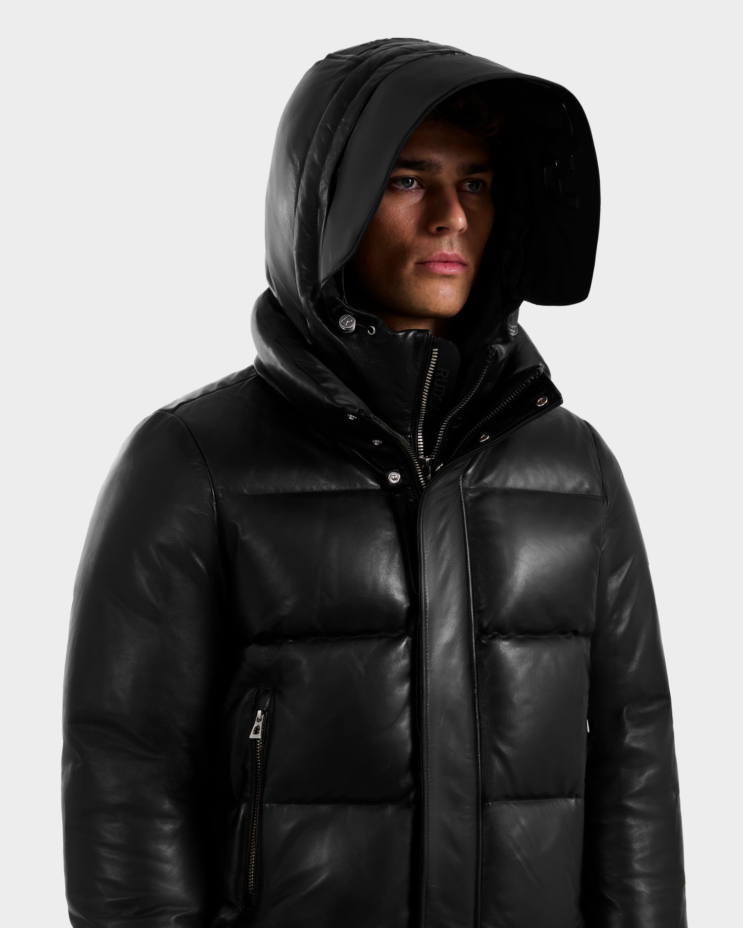 Someone in a shiny black Rudsak JORDY X - 6125694 unisex jacket with a large hood up, looking slightly to the side against a plain light background.