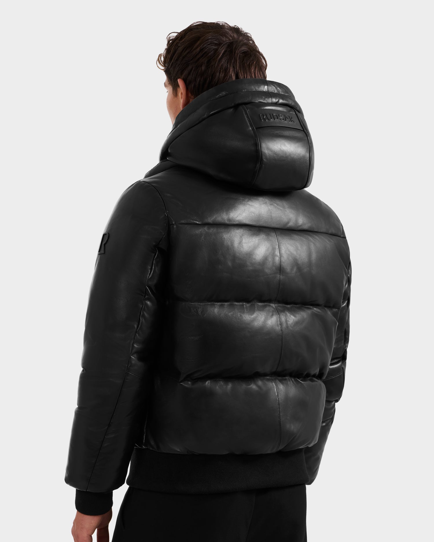 A person is shown from the back wearing the Rudsak JORDY X - 6125694 Black unisex luxe leather puffer jacket with a hood, standing against a plain light gray background.