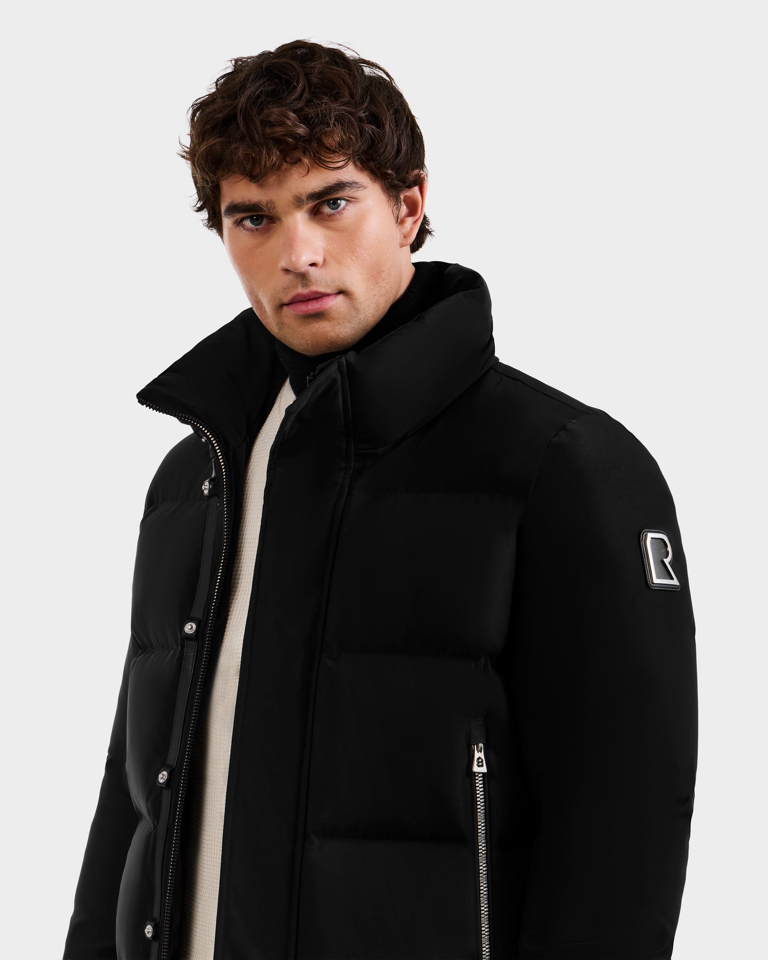A man with short, curly brown hair wears the Rudsak JORDY - 6125692 Black puffer jacket featuring a high collar, zippered pockets, and a sleeve patch. He looks directly at the camera against a plain light background.