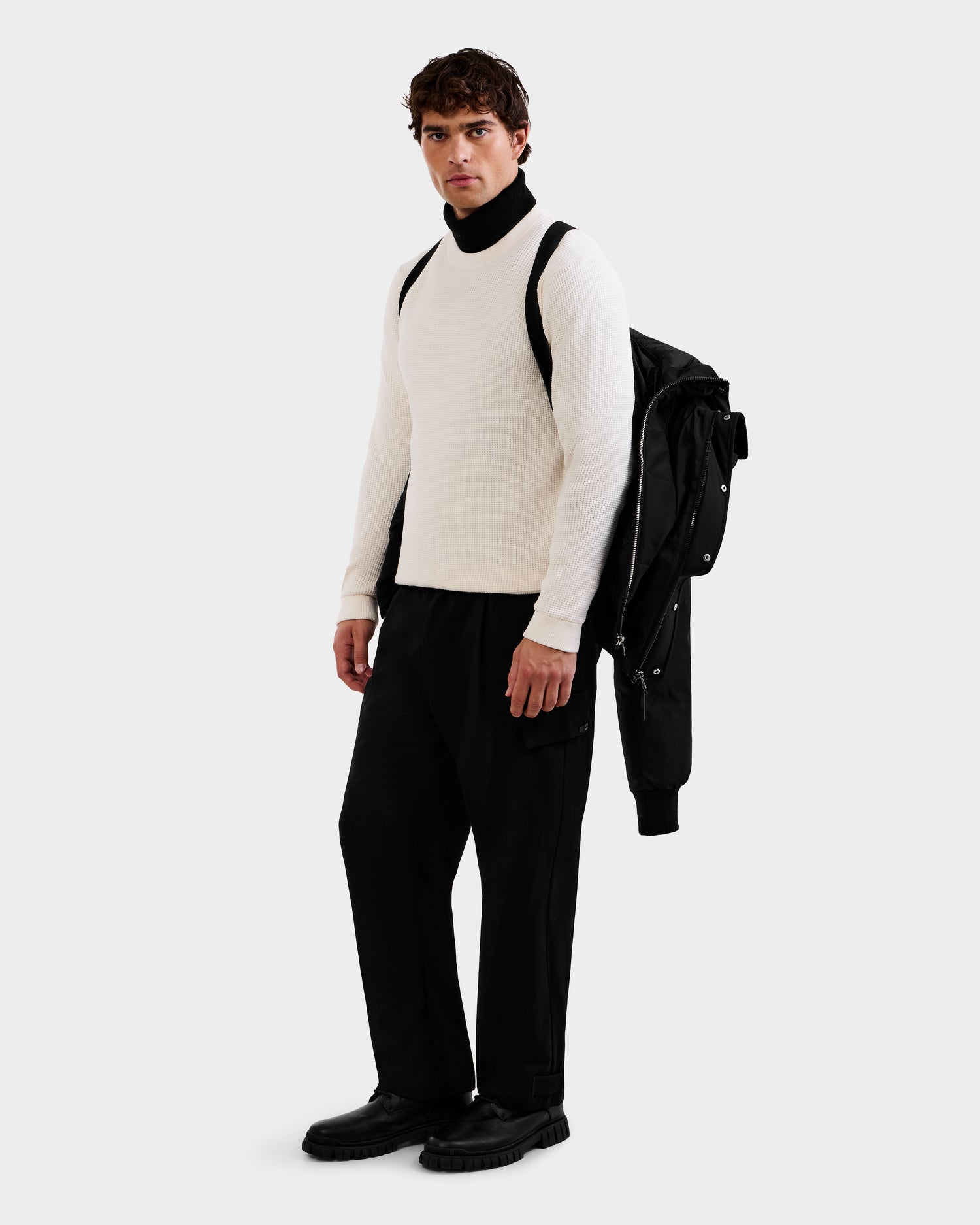 A man faces forward, wearing the Rudsak JORDY - 6125692 Black jacket draped over one shoulder and carrying a black backpack. He pairs it with a white sweater, black turtleneck, black pants, and black shoes against a plain white background.