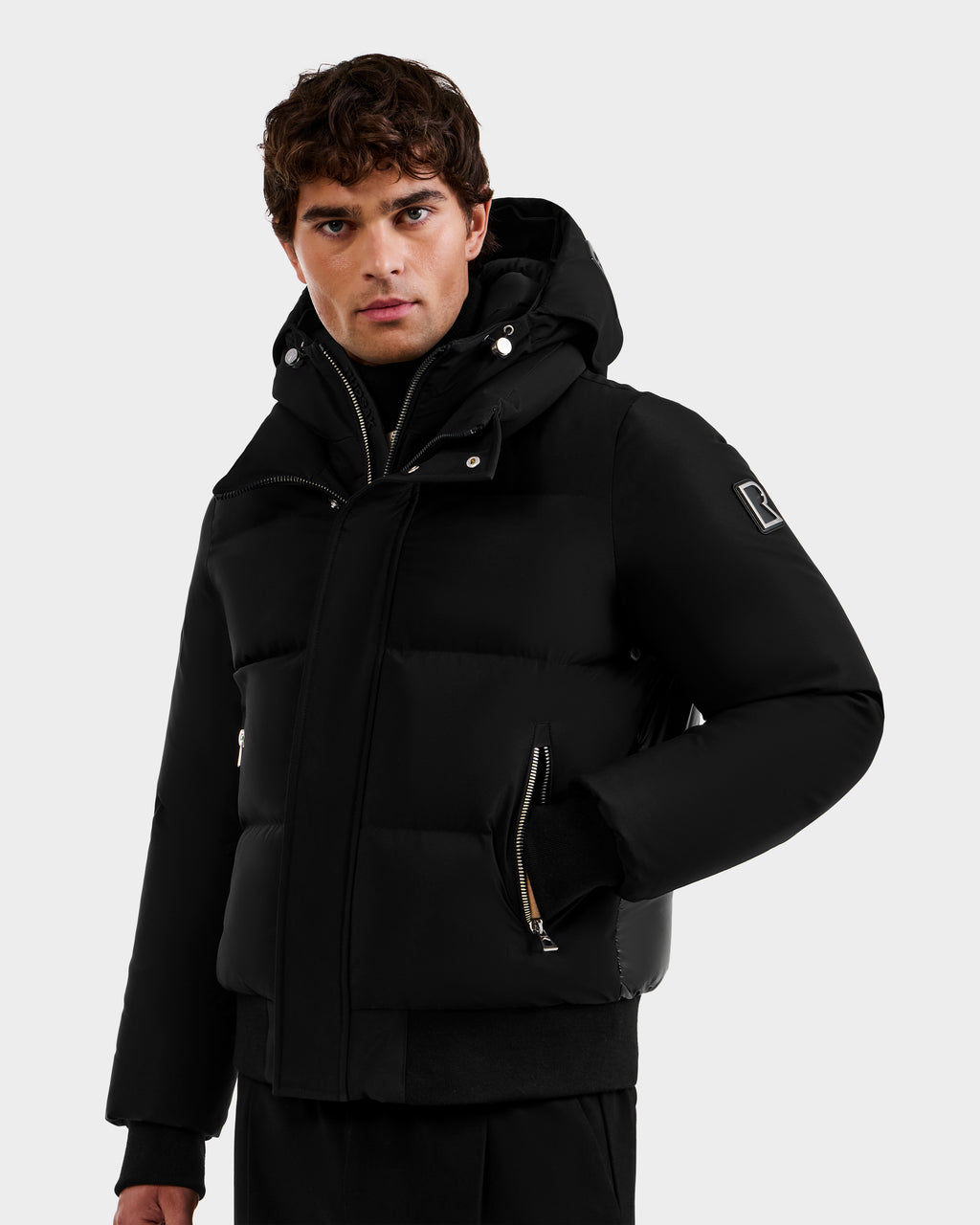A man with short, curly brown hair wears the Rudsak JORDY - 6125692 Black men's classic bomber puffer jacket with zippered pockets and a sleeve patch, featuring RDS down insulation, standing against a light gray background.