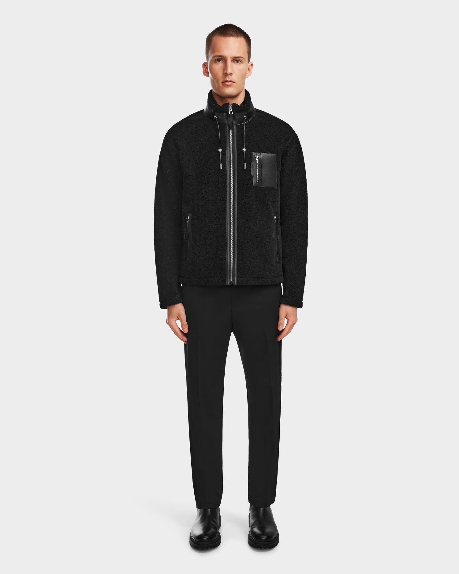 A man stands facing forward against a plain white background, wearing the Rudsak JOSEPH - 6524535 Black zip-up jacket paired with black pants and black shoes.