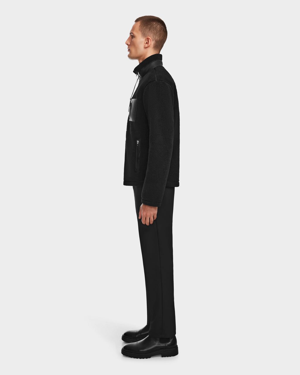 A man in profile facing left wears the Rudsak JOSEPH - 6524535 Black vegan leather jacket, black pants, and black boots against a plain white background.